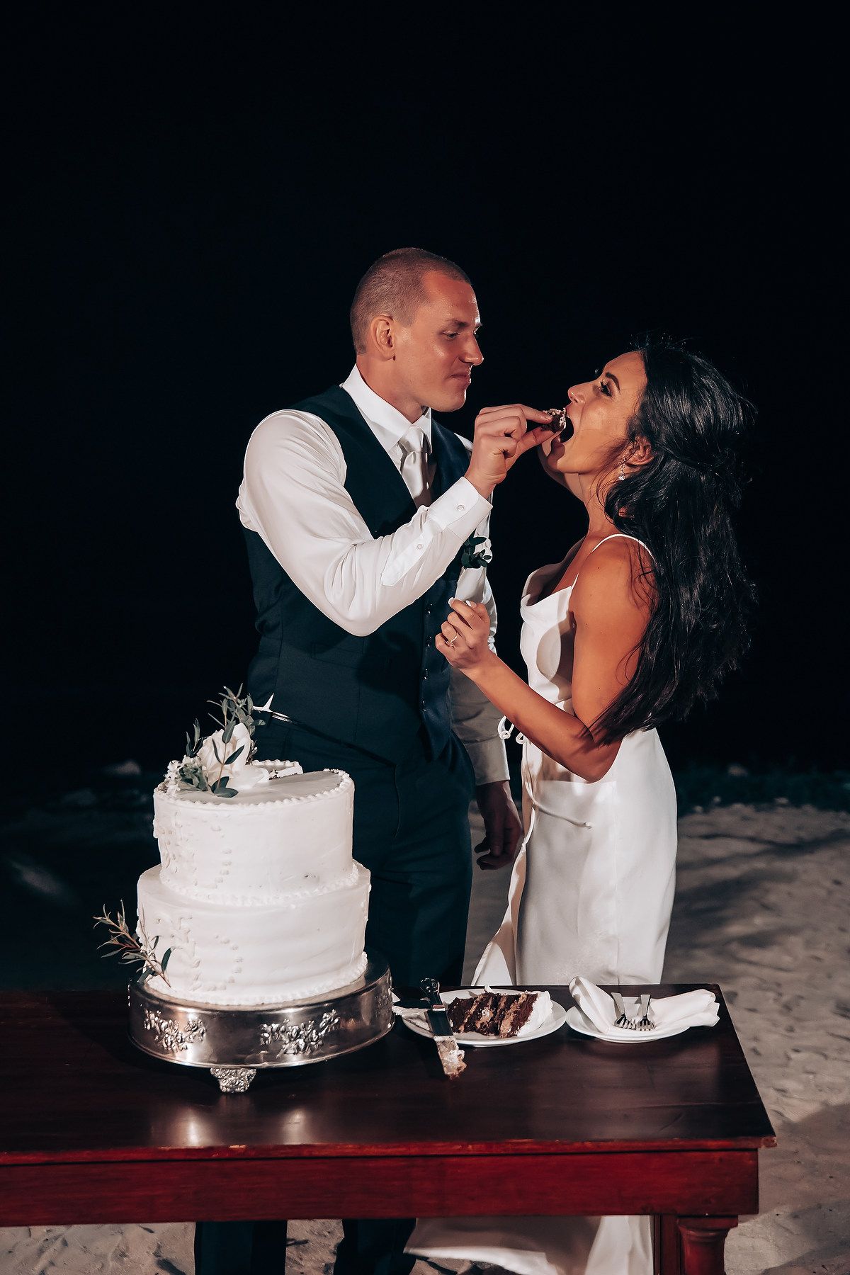 Newlyweds sharing cake feeding moment at their wedding reception against dark background.