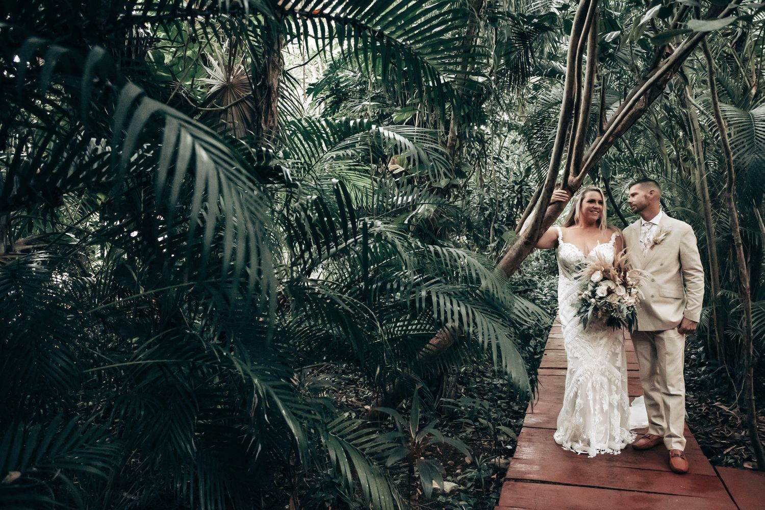A bride and groom stand together on a wooden bridge surrounded by dense tropical jungle foliage.