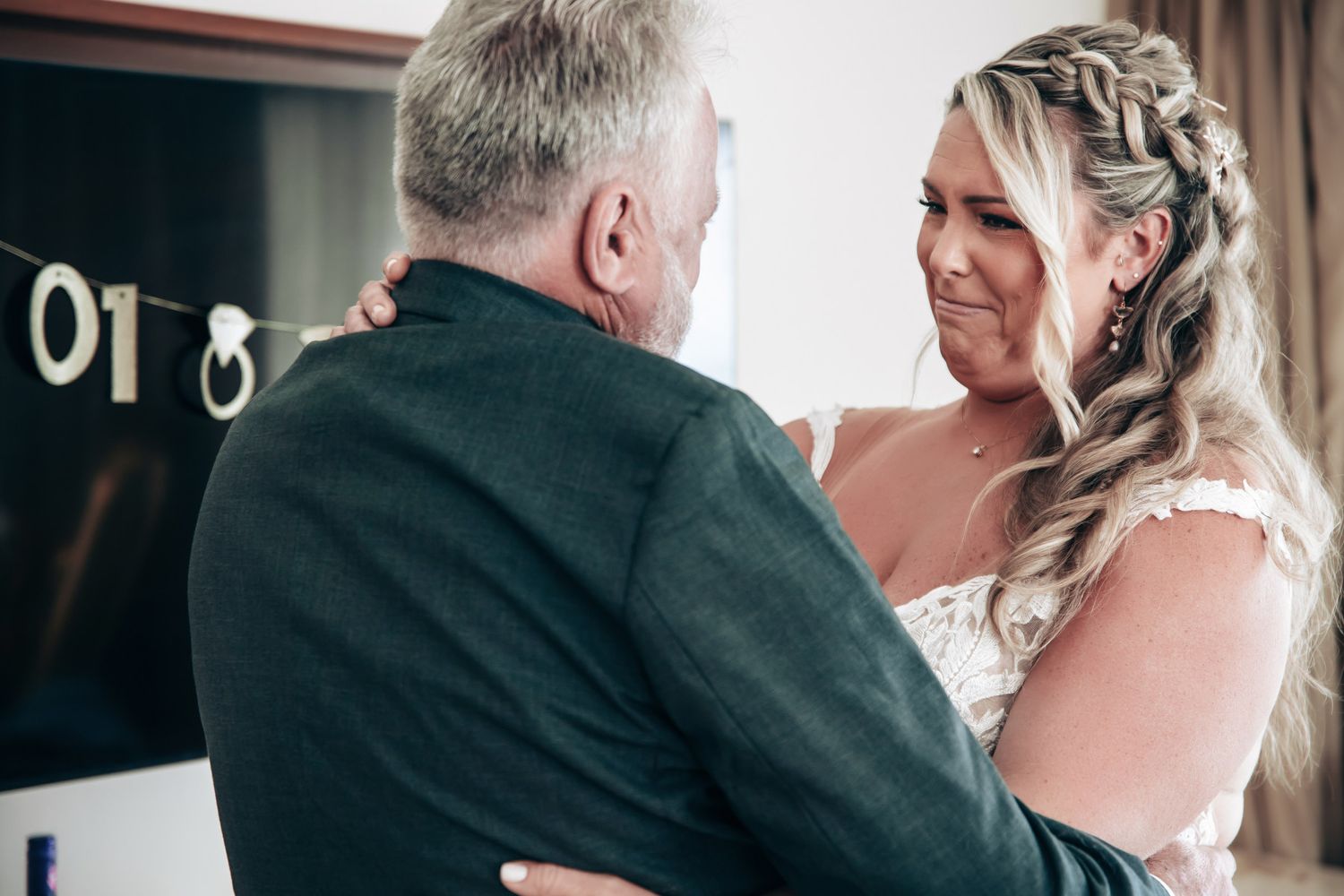 Emotional bride in lace wedding dress embracing older man, likely father, with tears of joy before wedding ceremony.