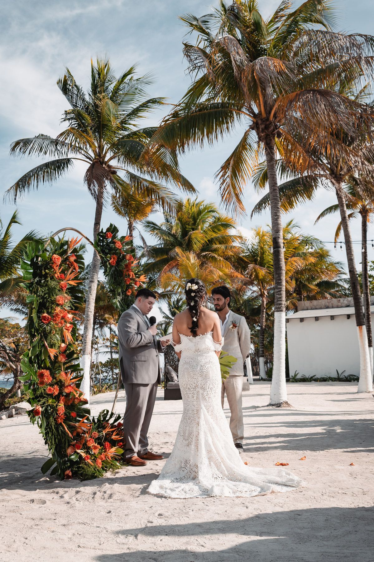 Bride and groom exchange vows under orange floral arch surrounded by palm trees on sandy beach.