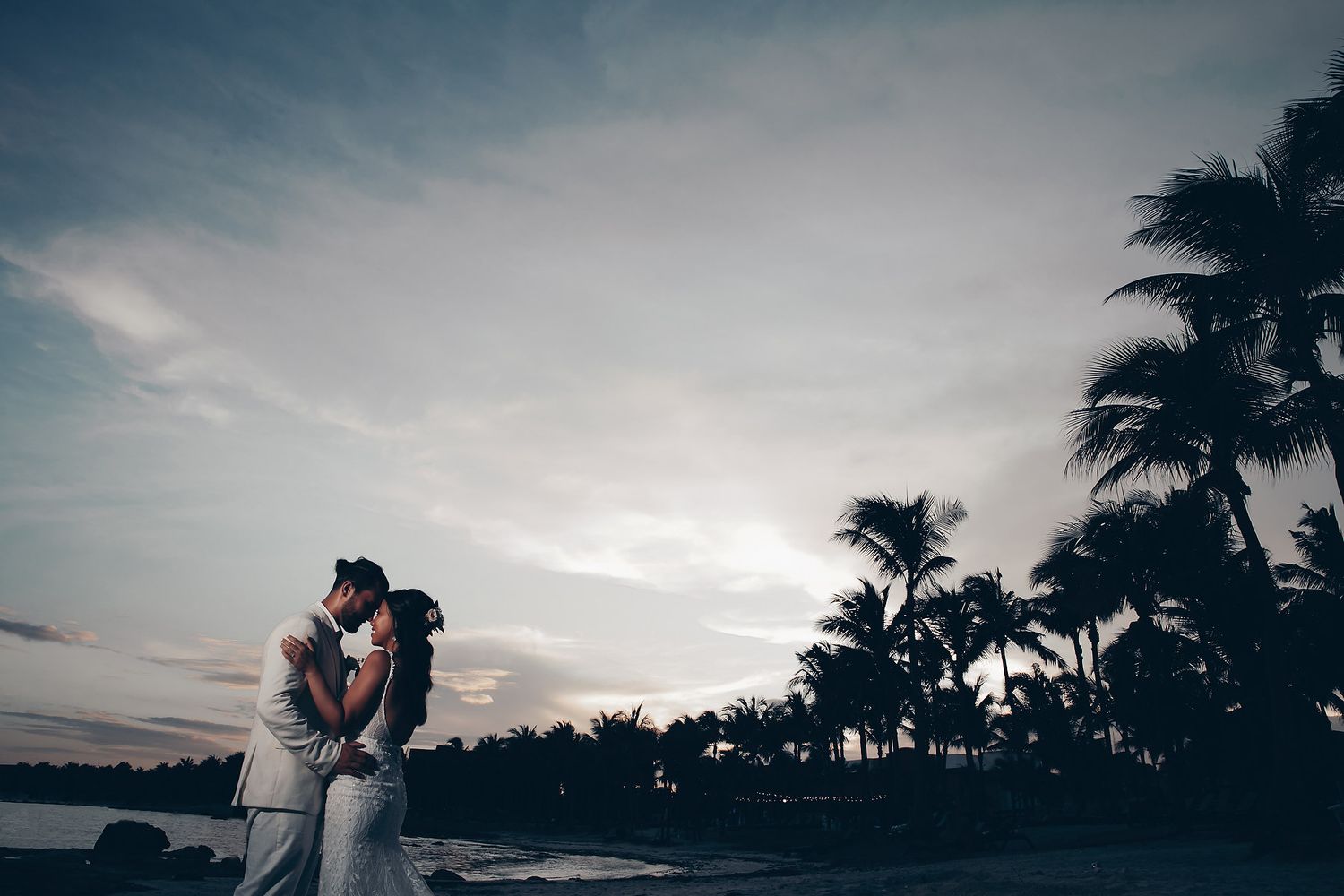 Silhouetted palm trees and dramatic sky create romantic backdrop for beach wedding photography.