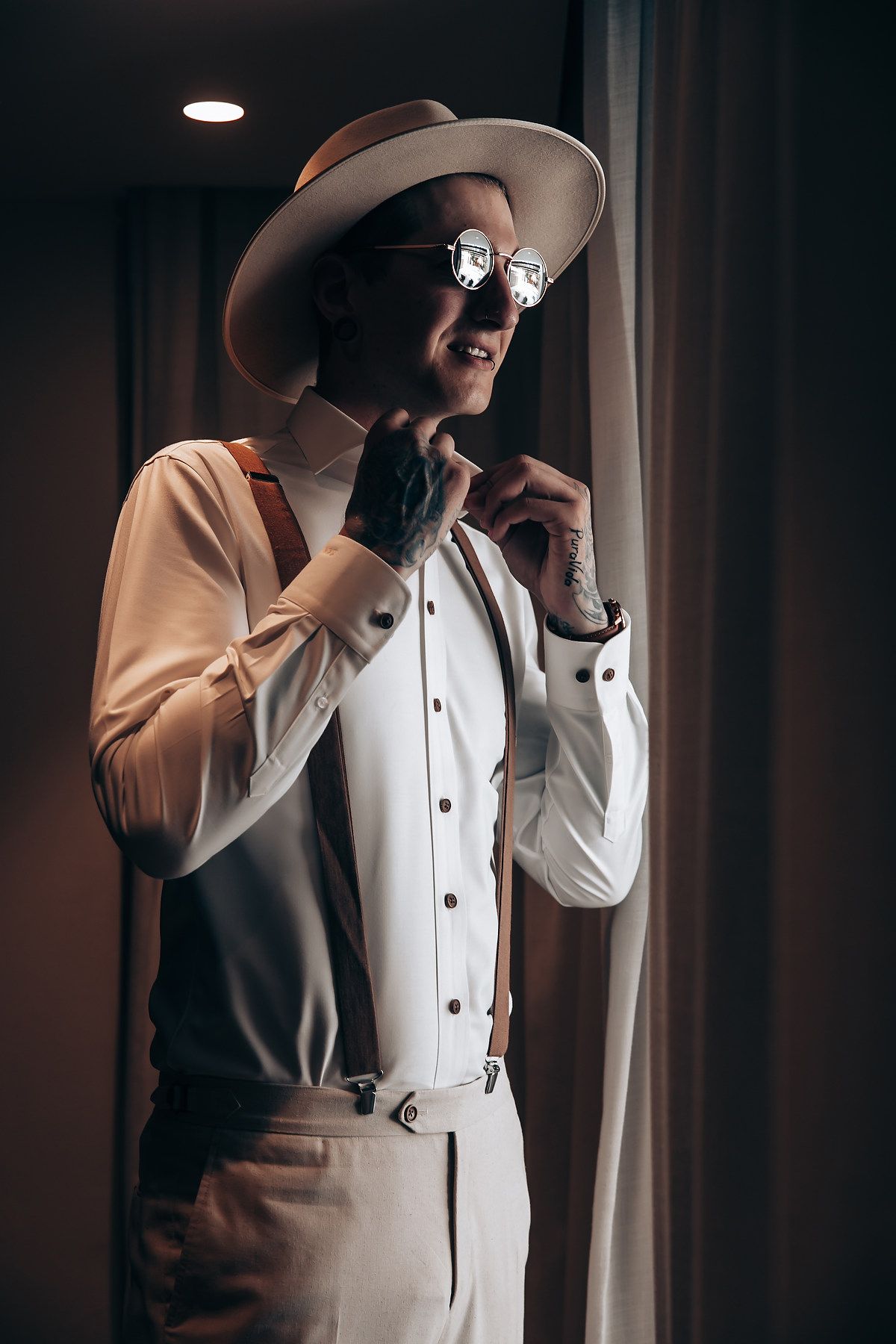 Stylish figure in vintage-inspired outfit with fedora hat, suspenders and round sunglasses poses near window in moody lighting.