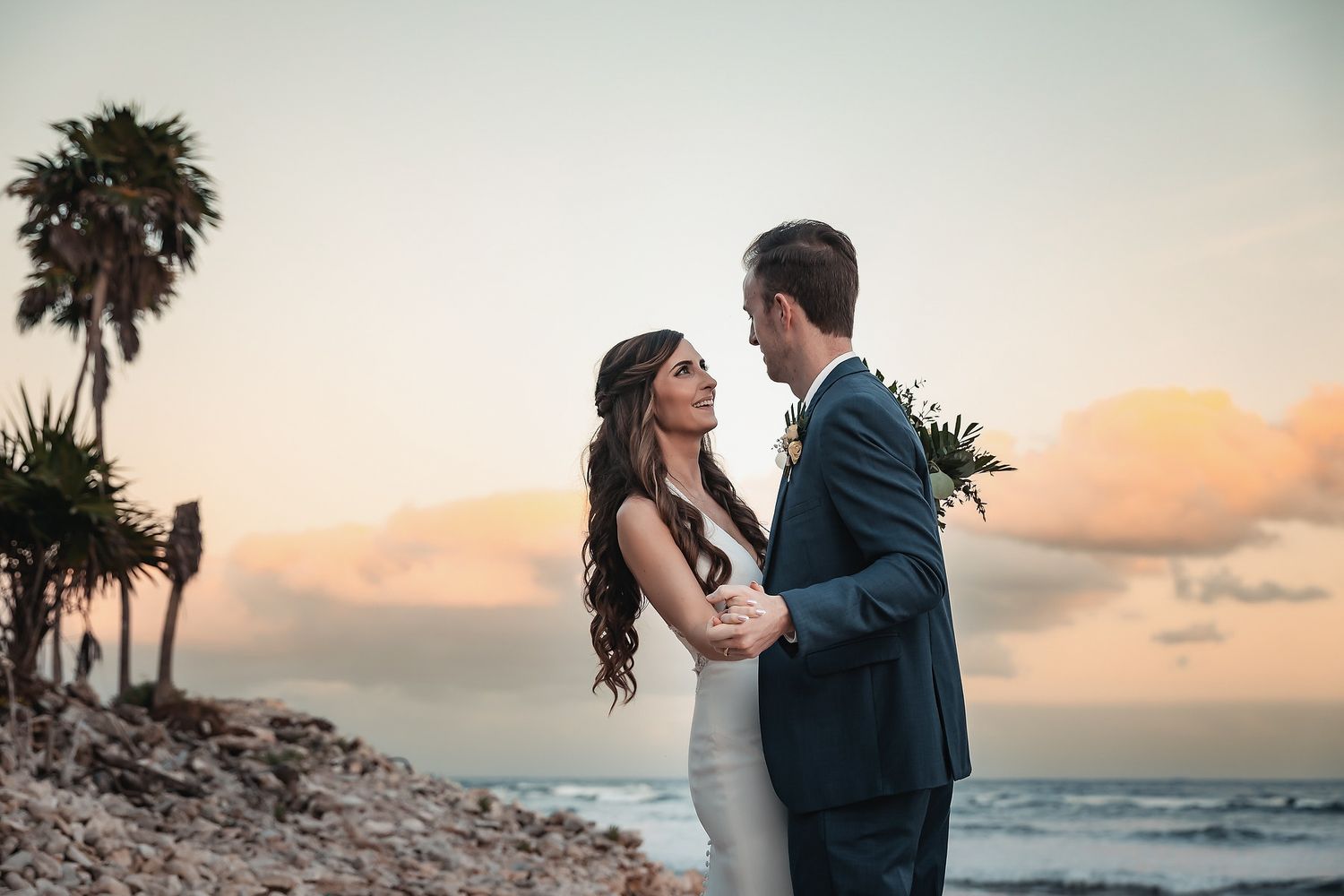 Bride and groom share a romantic moment on a rocky beach at sunset with palm trees behind them.