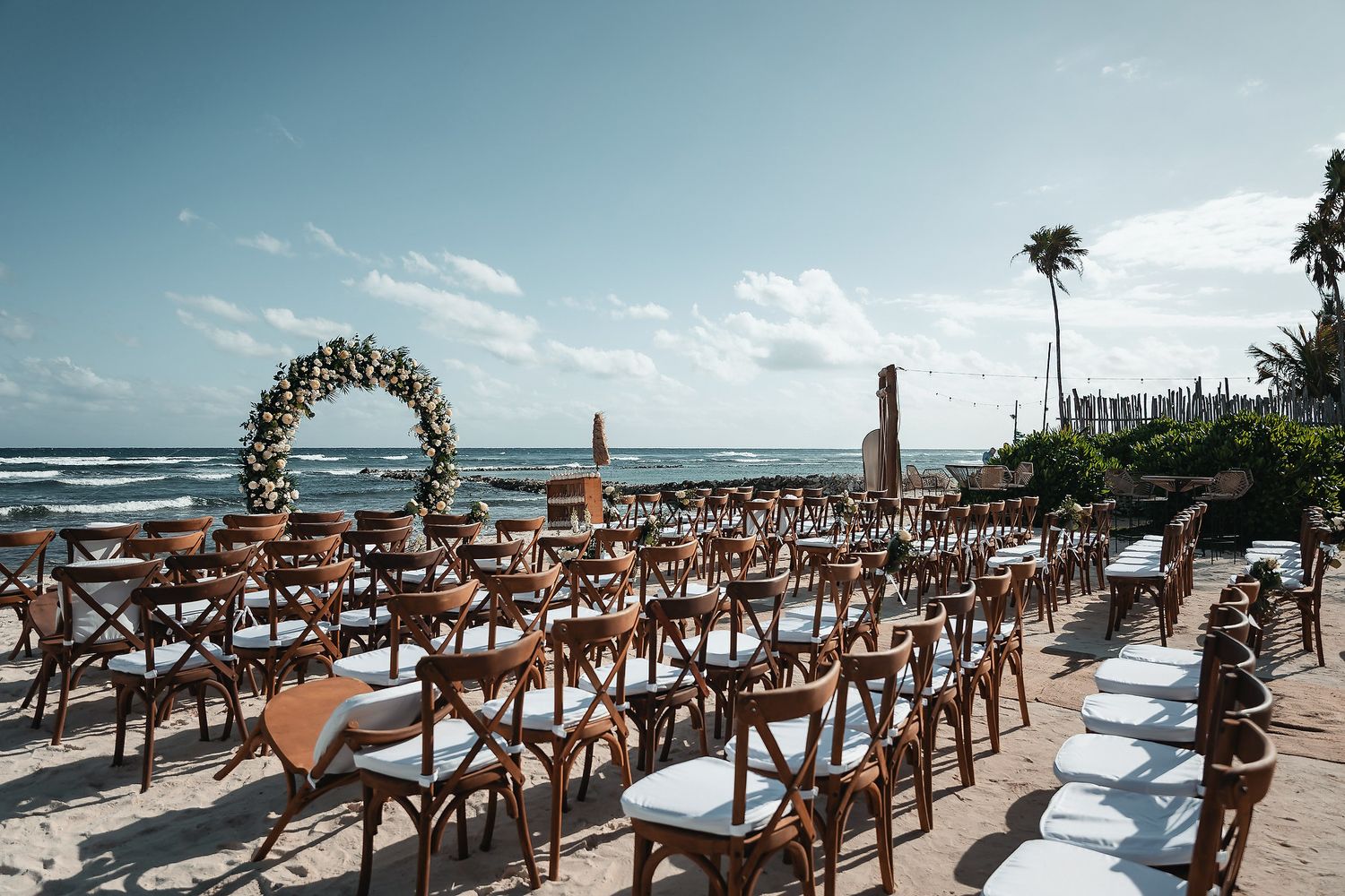 Beach wedding venue with wooden cross-back chairs, floral arch, and ocean backdrop under blue sky.