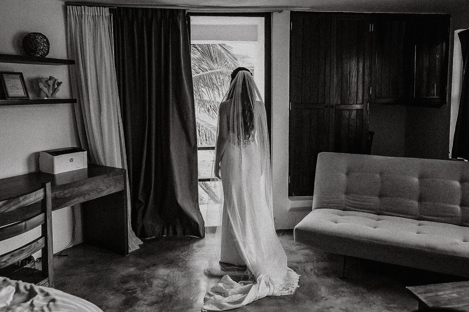 Black and white photo of a bride in wedding gown and veil standing in a doorway with natural light.