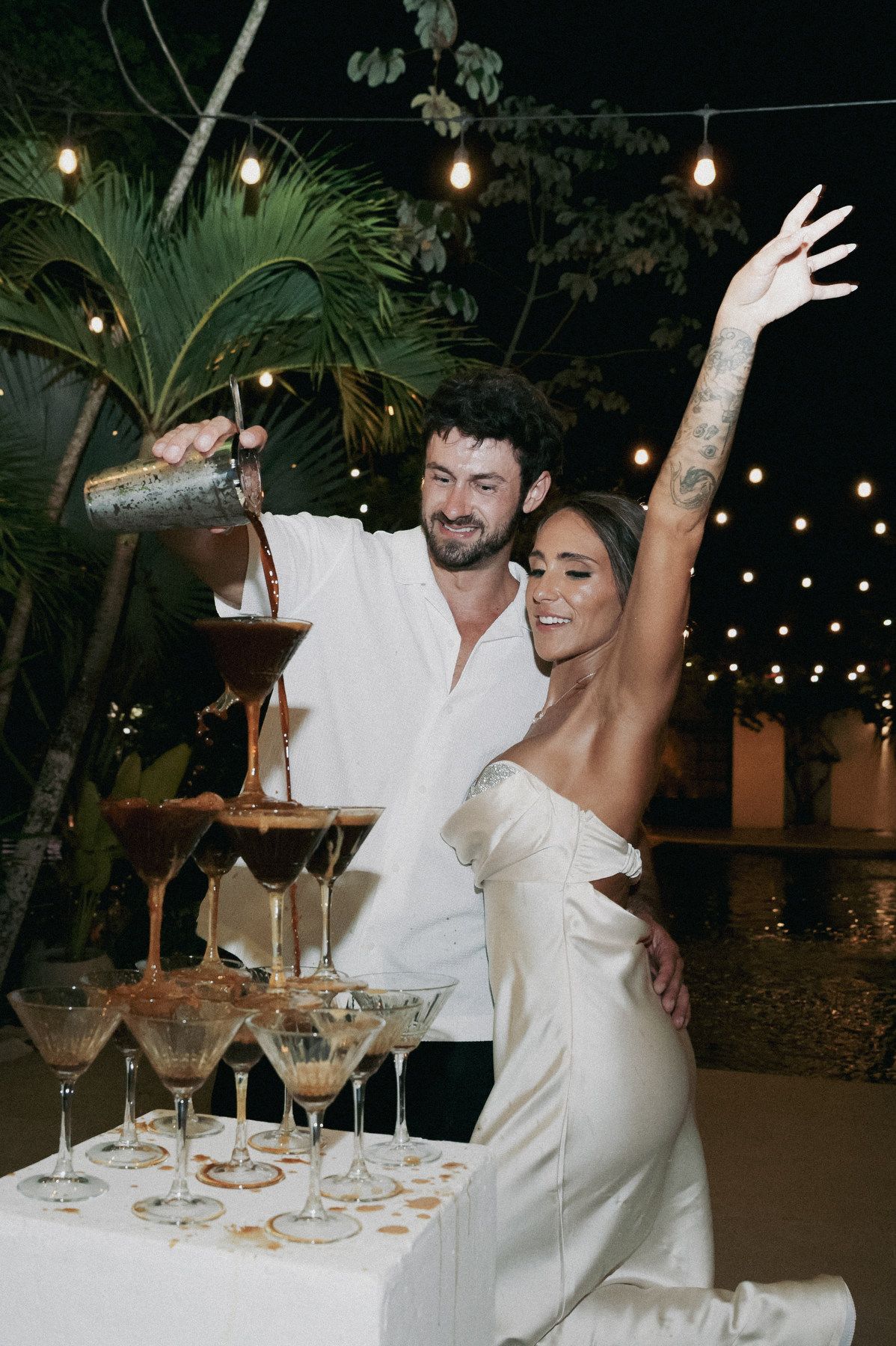 Couple celebrating at wedding espresso martini tower, bride in white dress raises arm while groom pours cocktails outdoors.