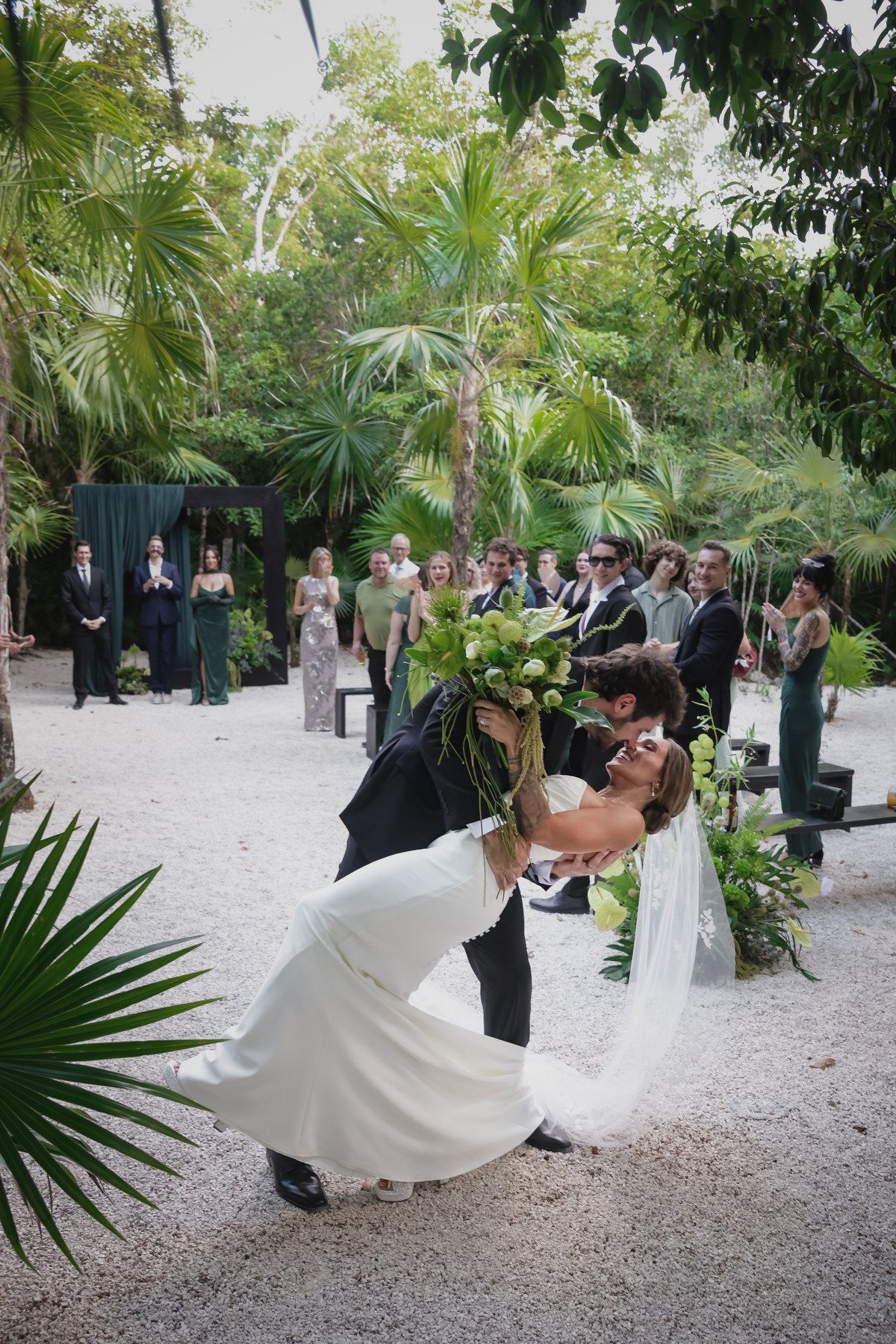 Groom dips bride for a kiss holding bouquet at lush tropical outdoor wedding ceremony with guests.