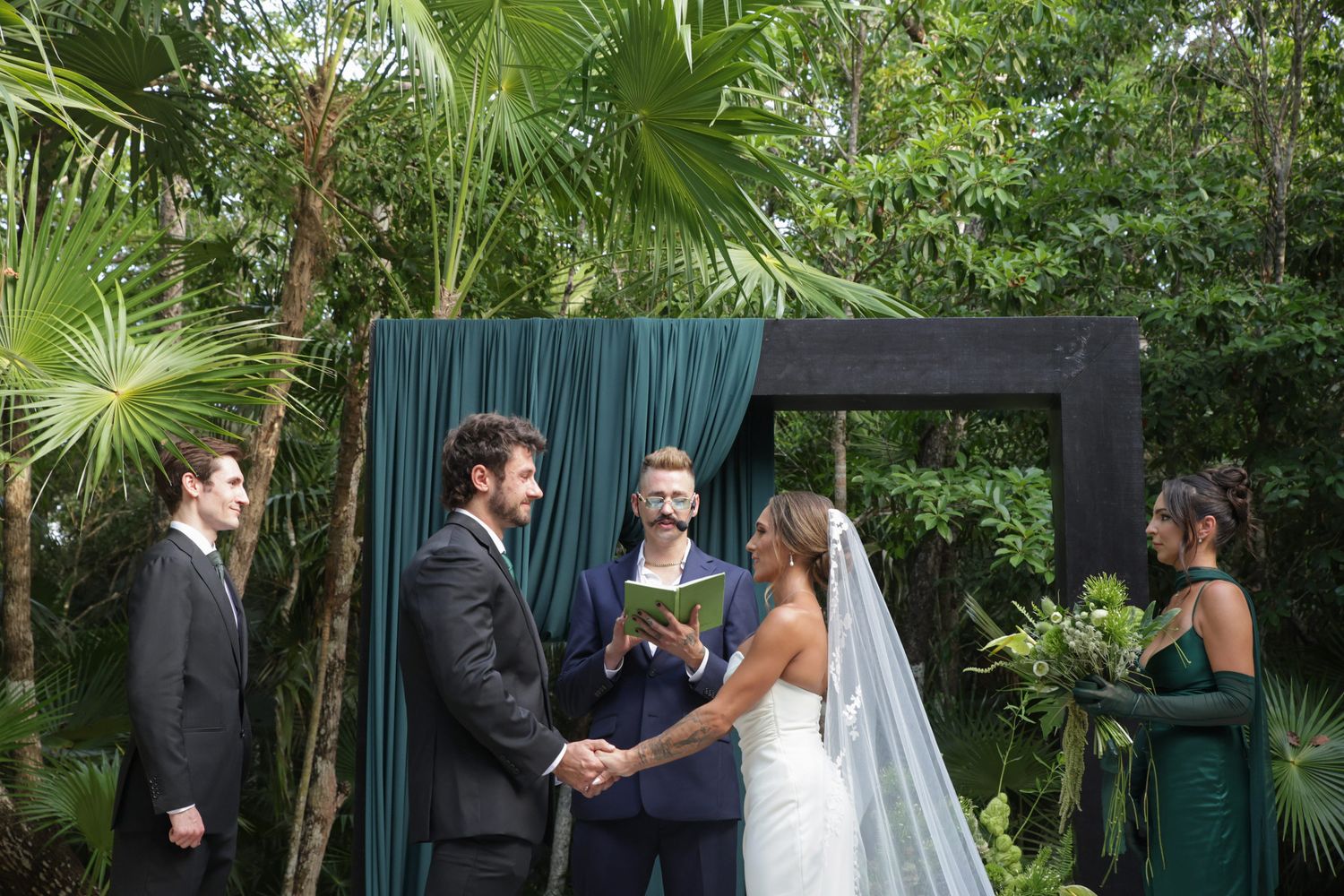 Outdoor wedding ceremony with couple exchanging vows before officiant, framed by dark arch with emerald draping amid lush tropical greenery.