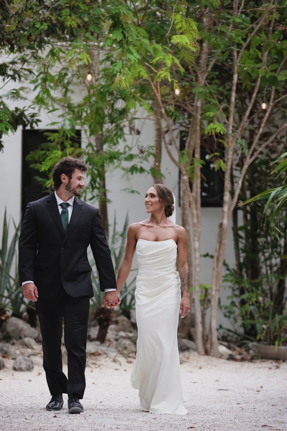 Bride and groom walk hand in hand in a tropical garden, smiling at each other on their wedding day.