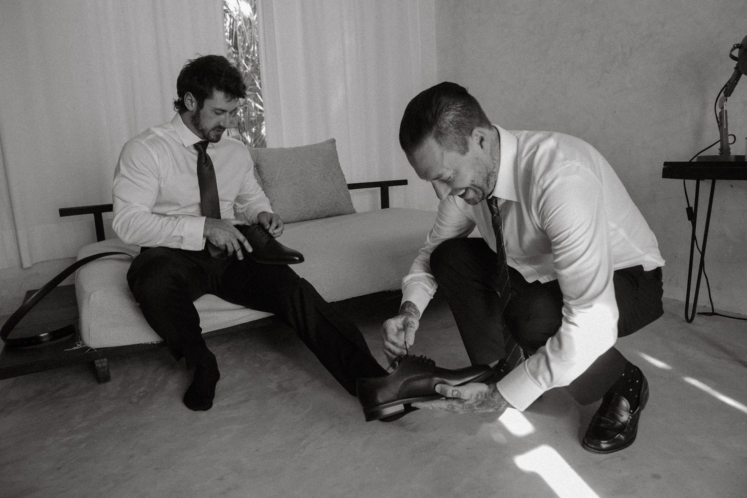 Groom getting shoes tied by groomsman in black and white wedding preparation photo.