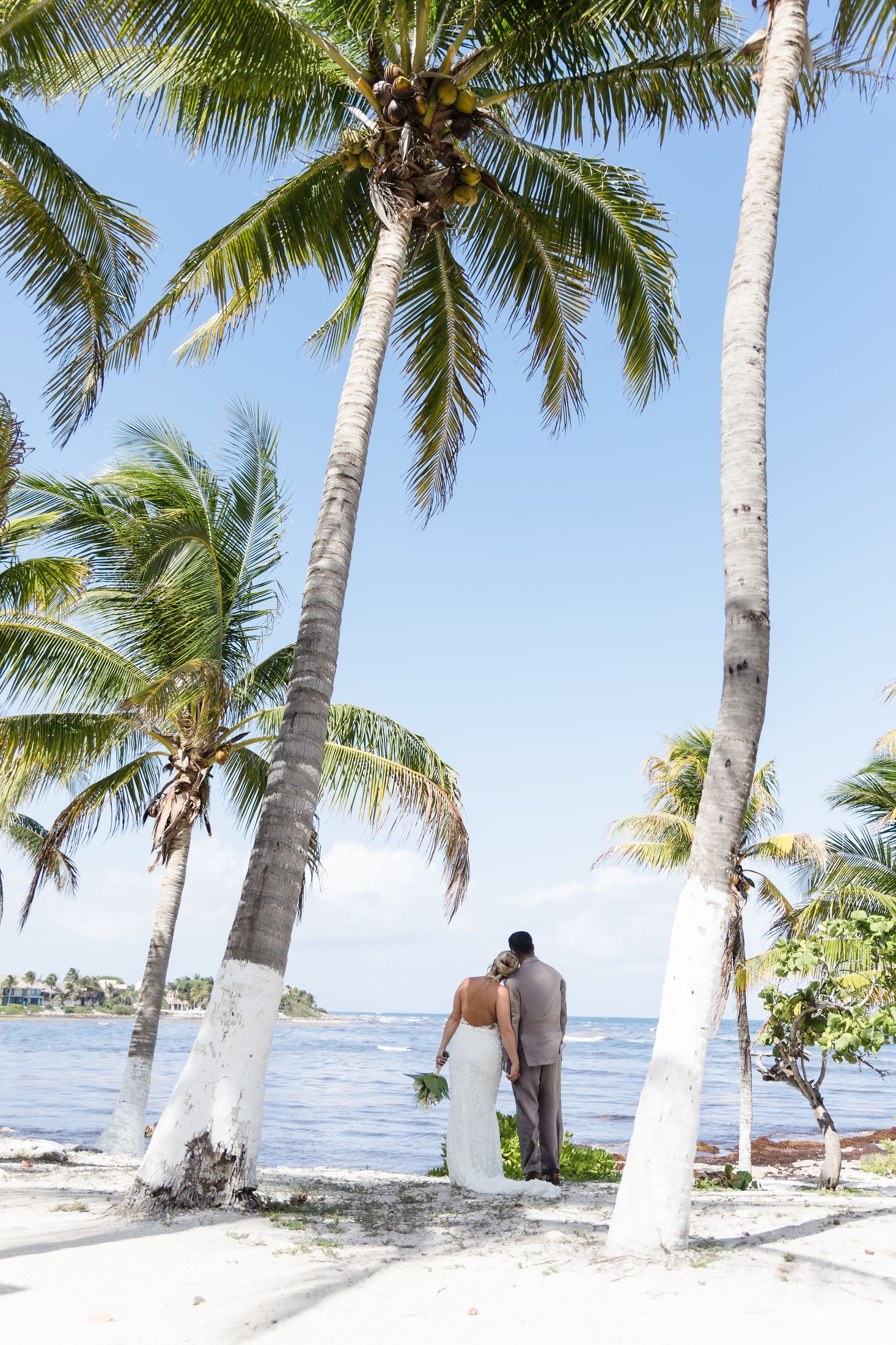Bride and groom stand among white palm trees on a tropical beach, gazing at the turquoise ocean on their wedding day.