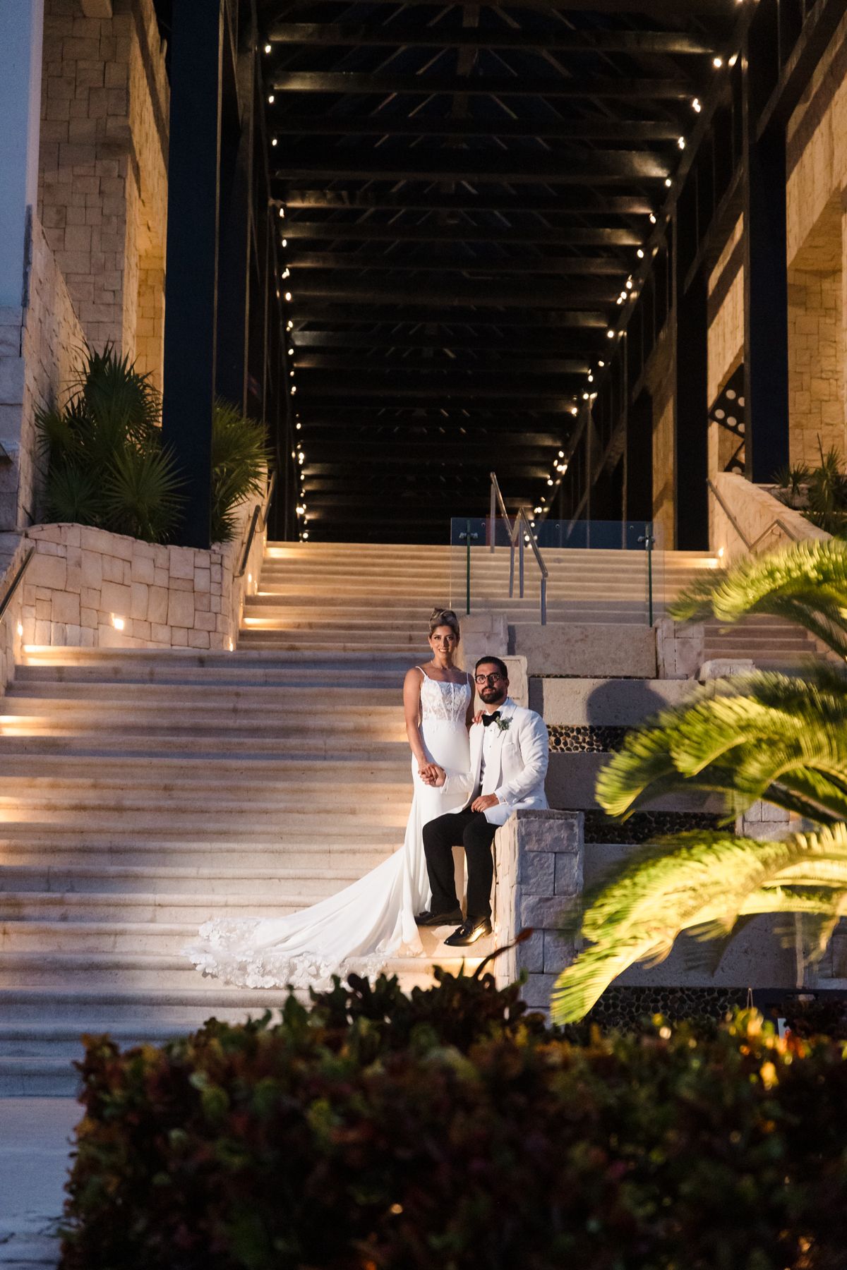 Bride and groom pose on grand marble steps under dramatic lit archway at elegant evening wedding venue.