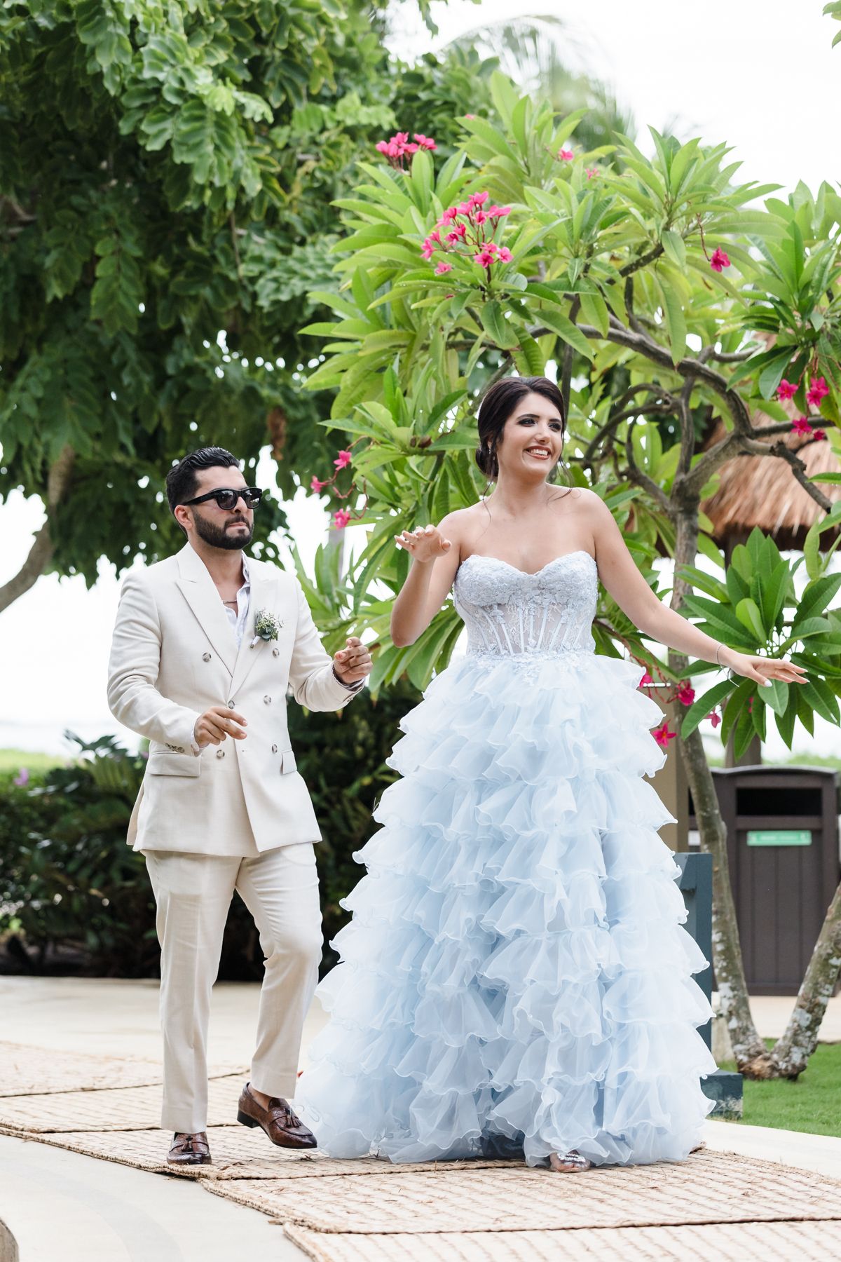 Bride in blue ruffled gown and groom in cream suit walk together outdoors near tropical trees with pink flowers.