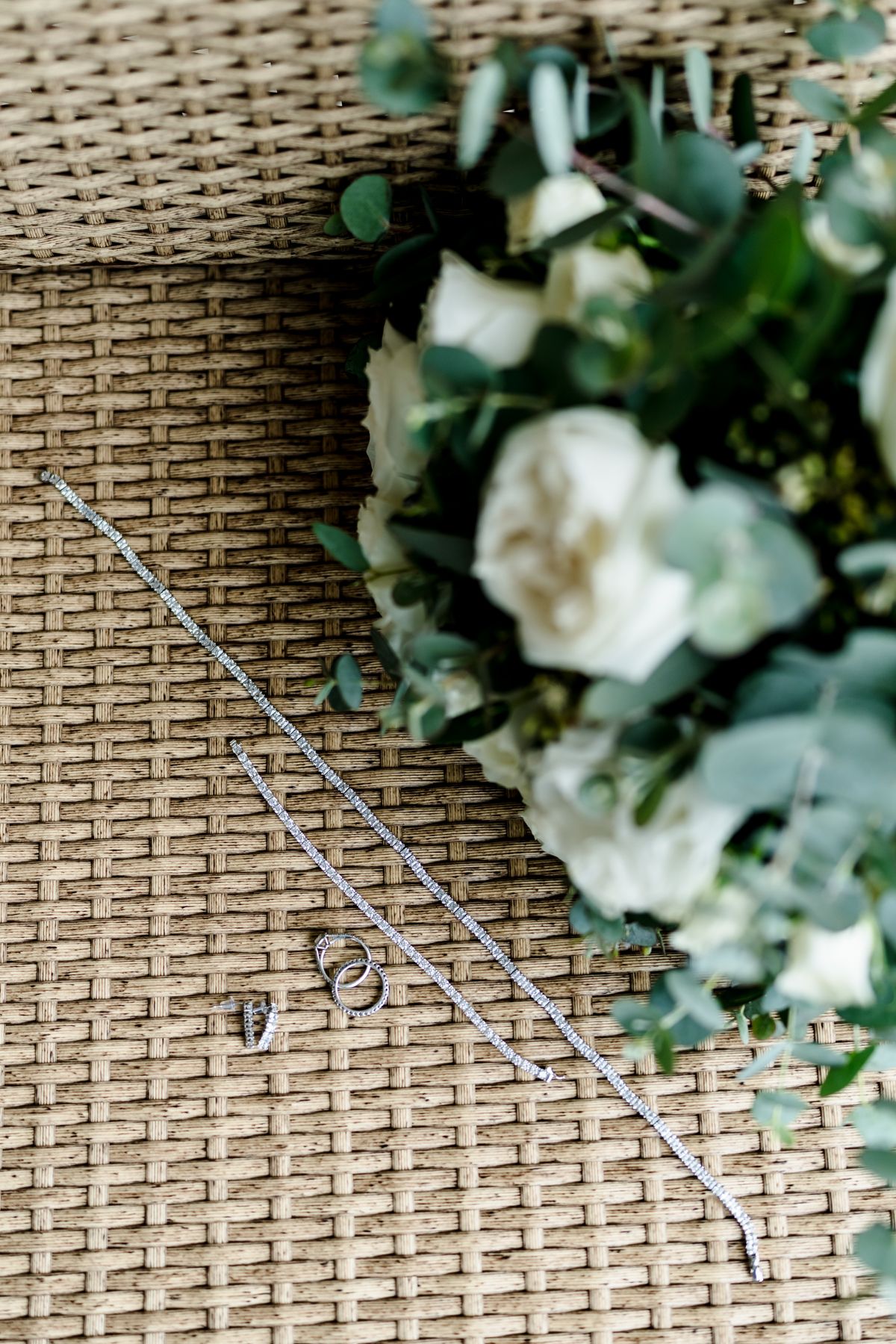 Wedding rings and silver bracelets arranged on woven texture beside a bouquet of white roses and eucalyptus greenery.