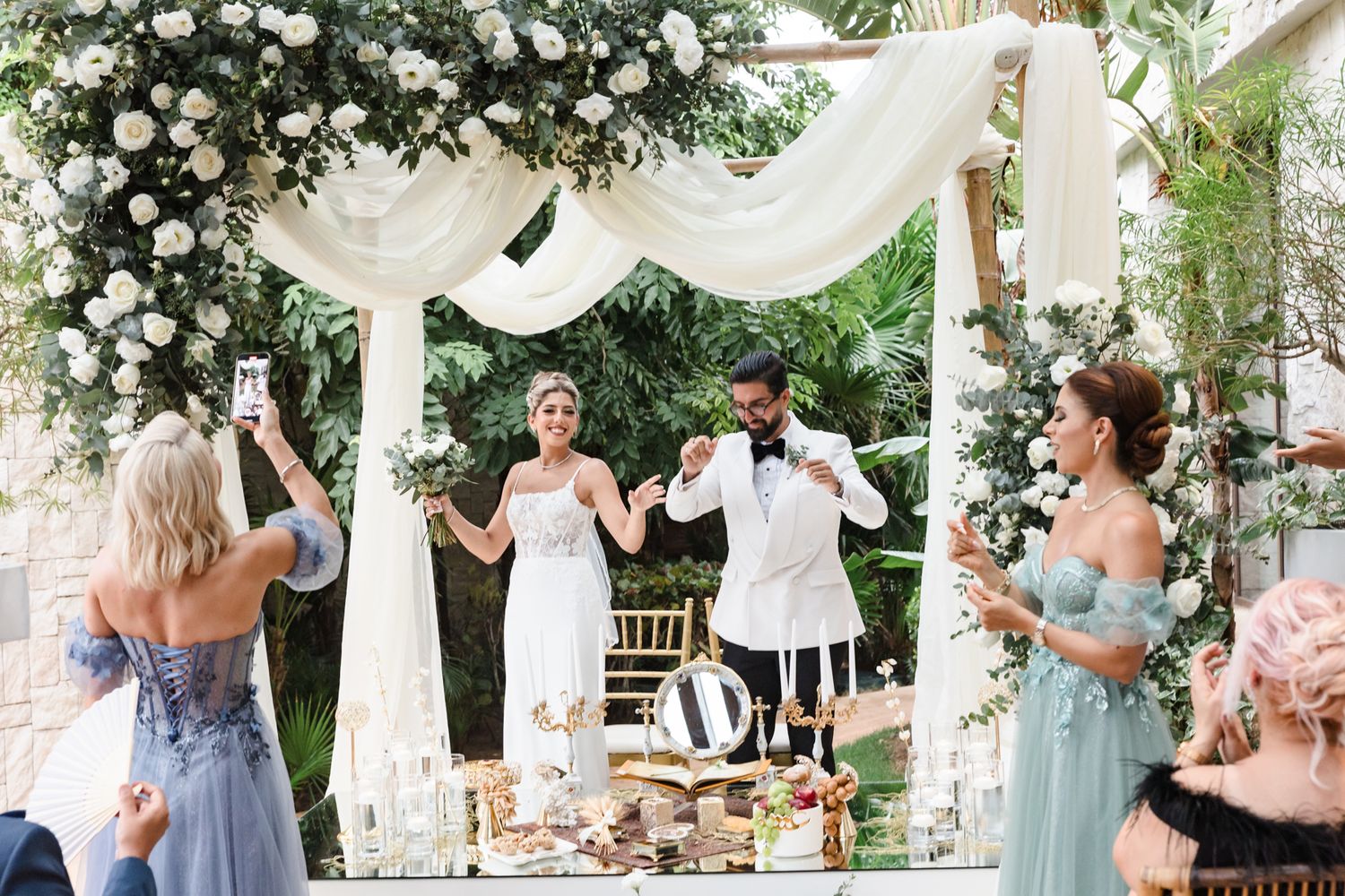 Bride and groom celebrate under floral arch with white draping at outdoor wedding reception with guests in blue dresses.