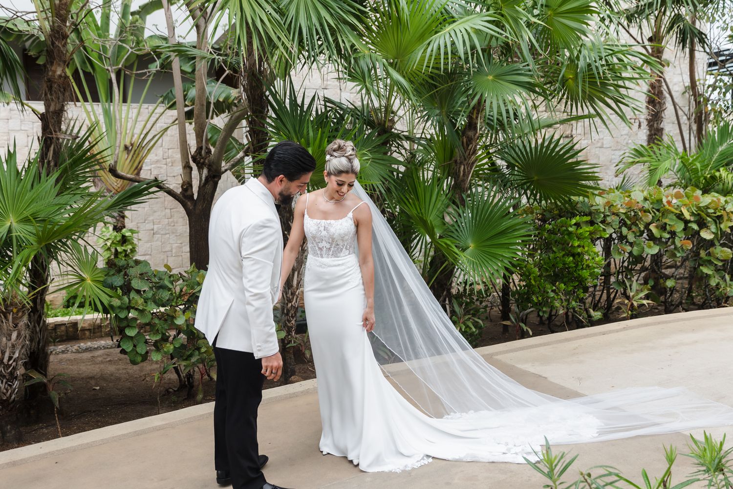 Bride in white gown and veil holds hands with groom in white jacket outdoors among tropical palm trees.