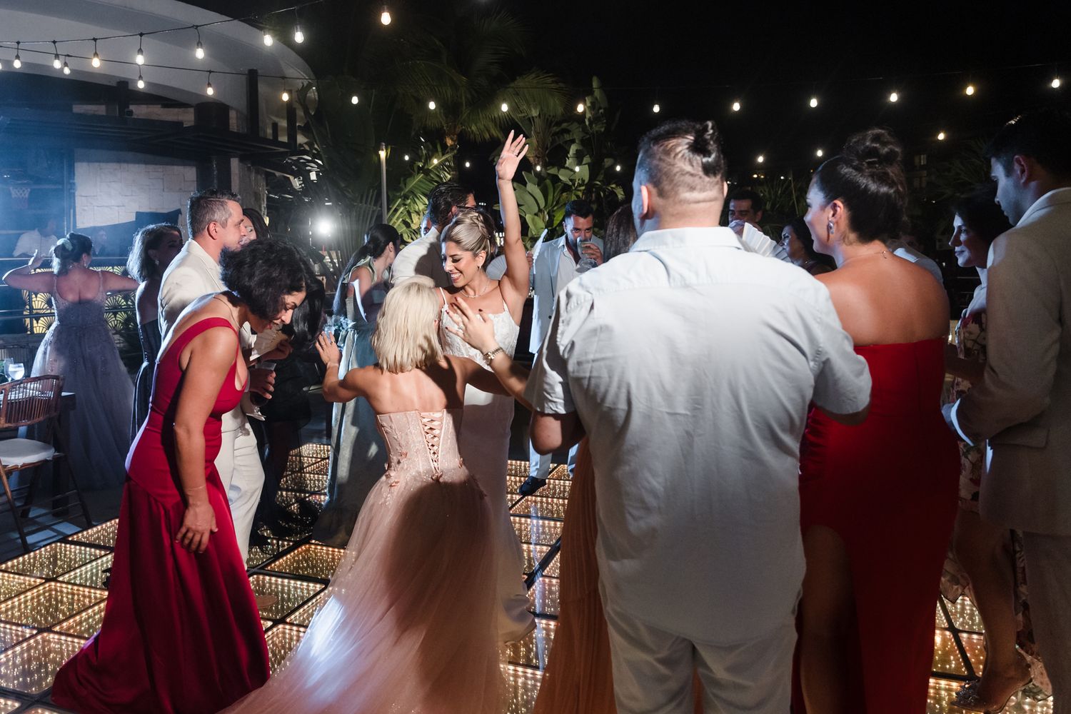 Bride dancing joyfully with wedding guests on illuminated dance floor at outdoor evening reception.