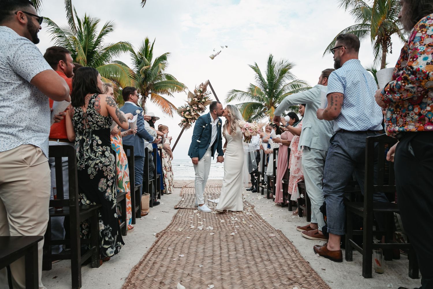 Newlyweds walk down aisle as guests celebrate with confetti at tropical beach wedding ceremony.
