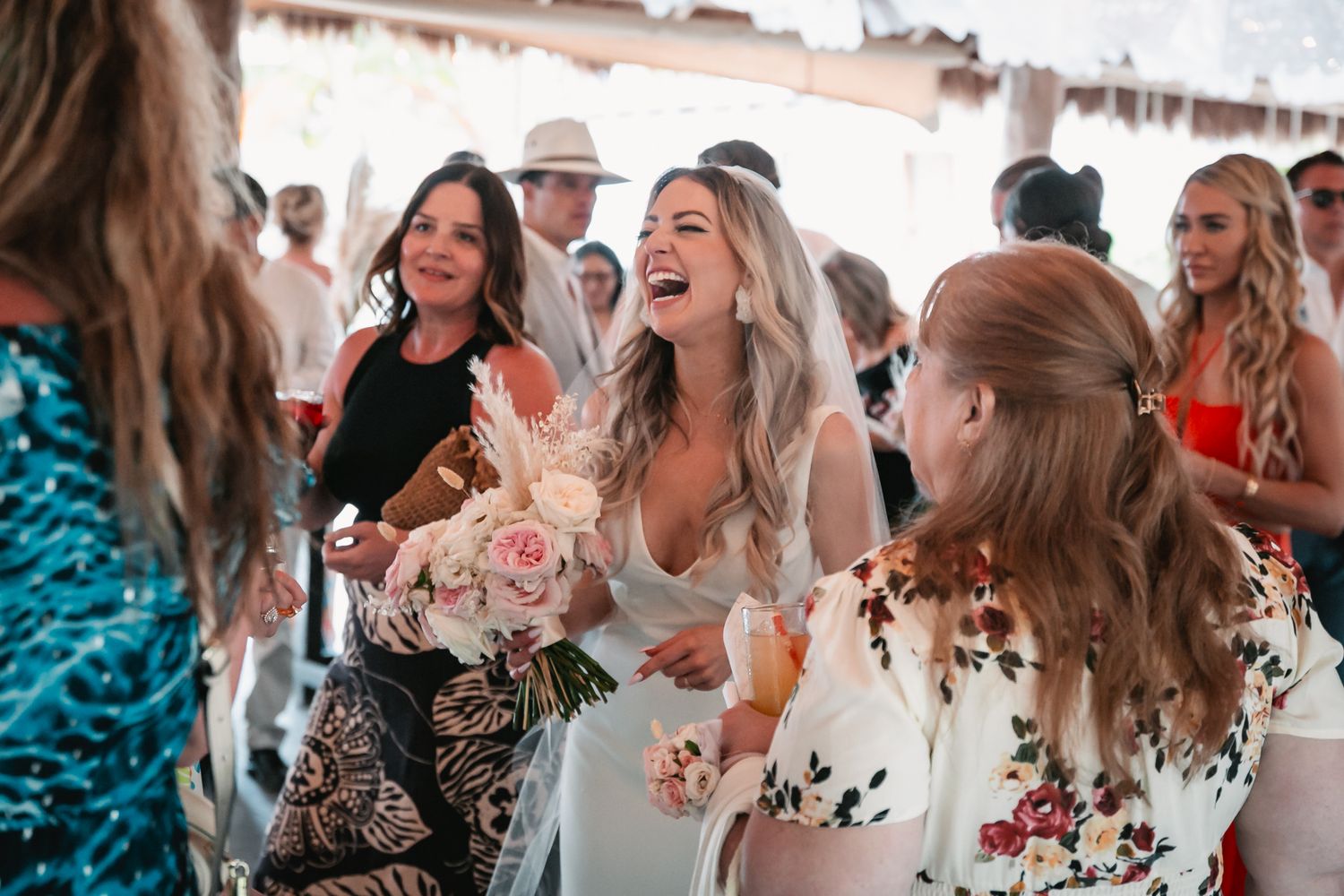 Laughing bride holding pink floral bouquet mingles with happy guests at a sunny outdoor wedding.