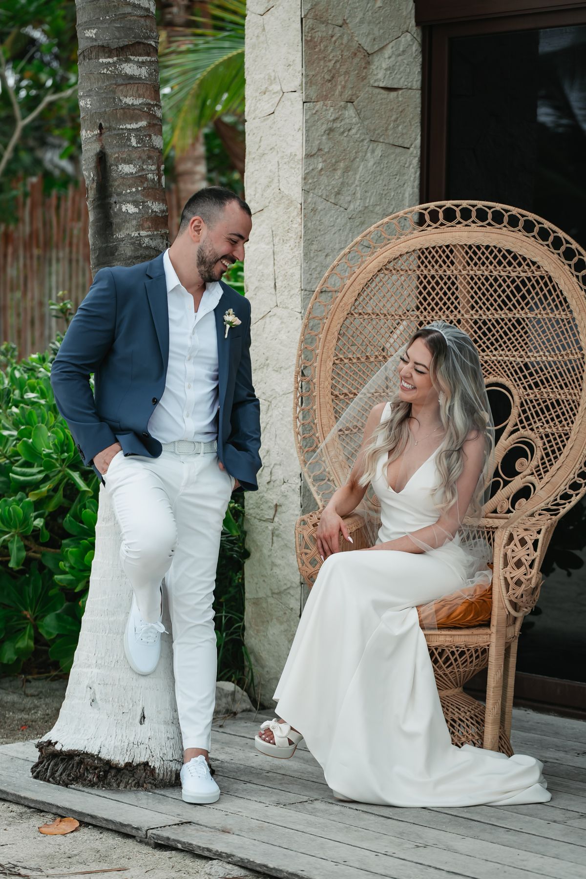 Smiling bride in white gown sits in peacock chair as groom in blue blazer leans on palm tree.