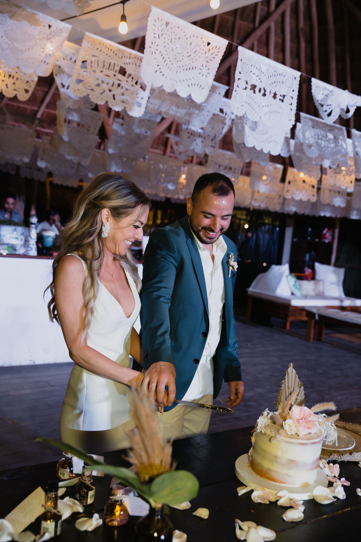 Bride and groom smiling while cutting a semi-naked wedding cake under white papel picado banners at reception.