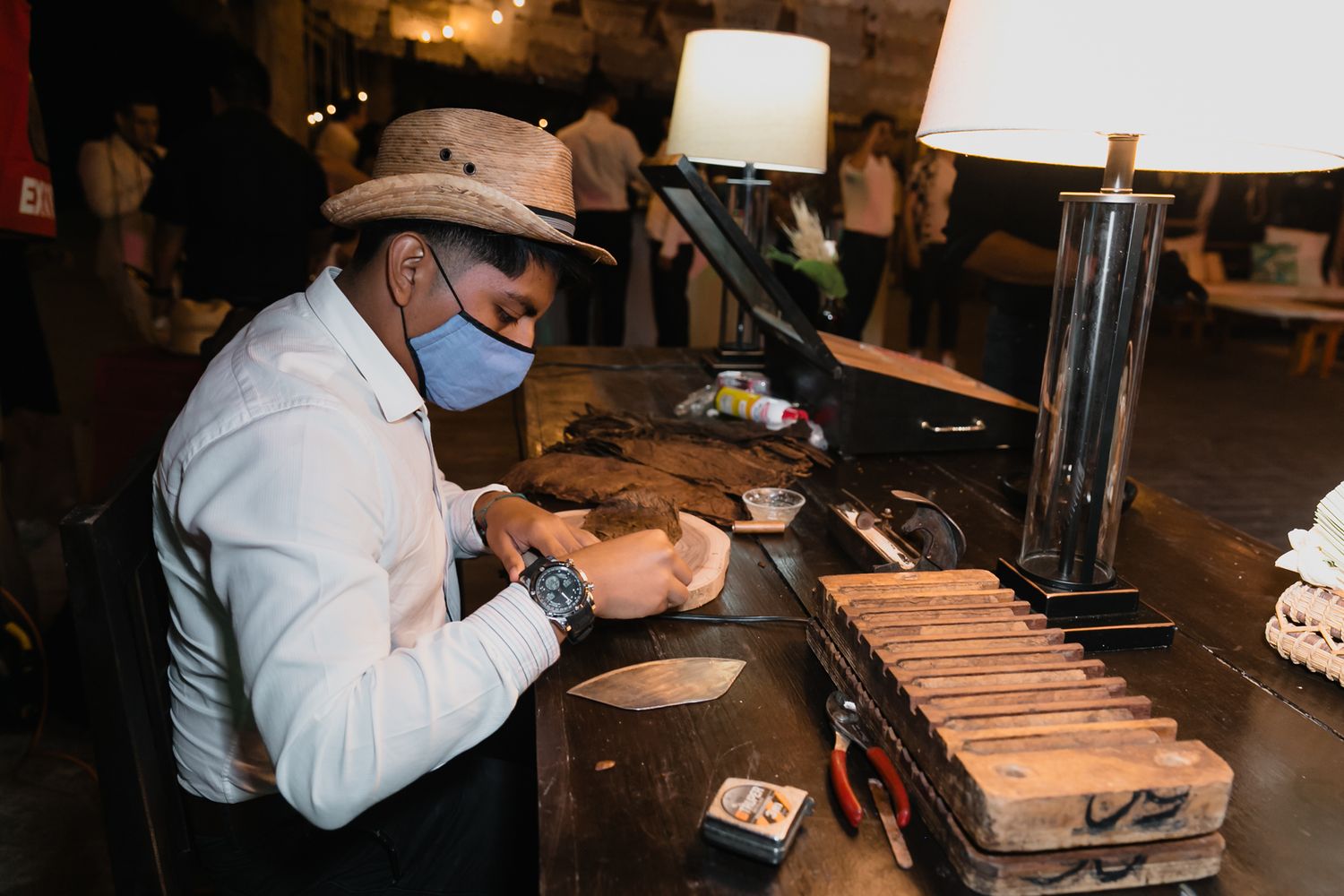 Masked cigar roller crafting cigars at wedding reception entertainment station for guests.