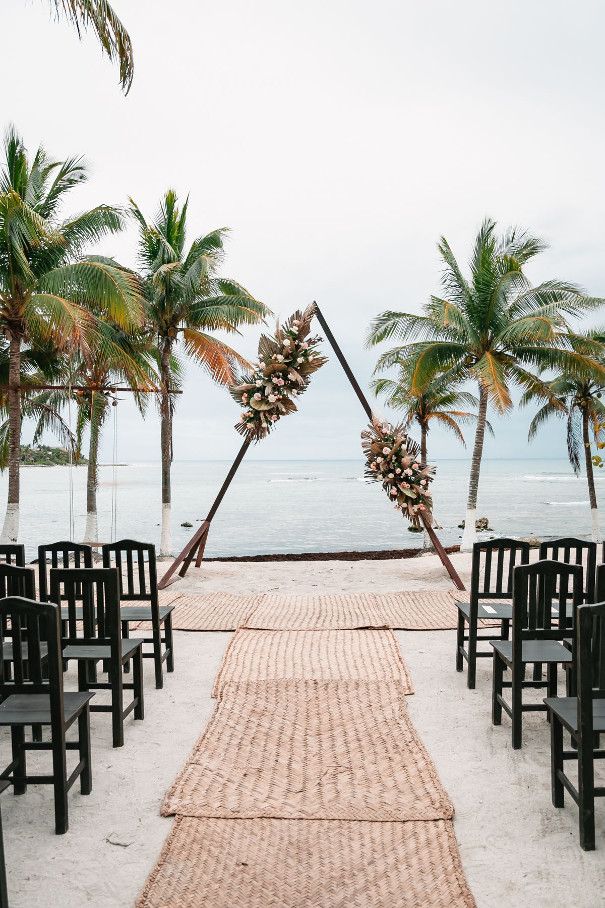 Beach wedding ceremony setup with triangular floral arch, woven aisle runner, and black chairs framed by palm trees and ocean.