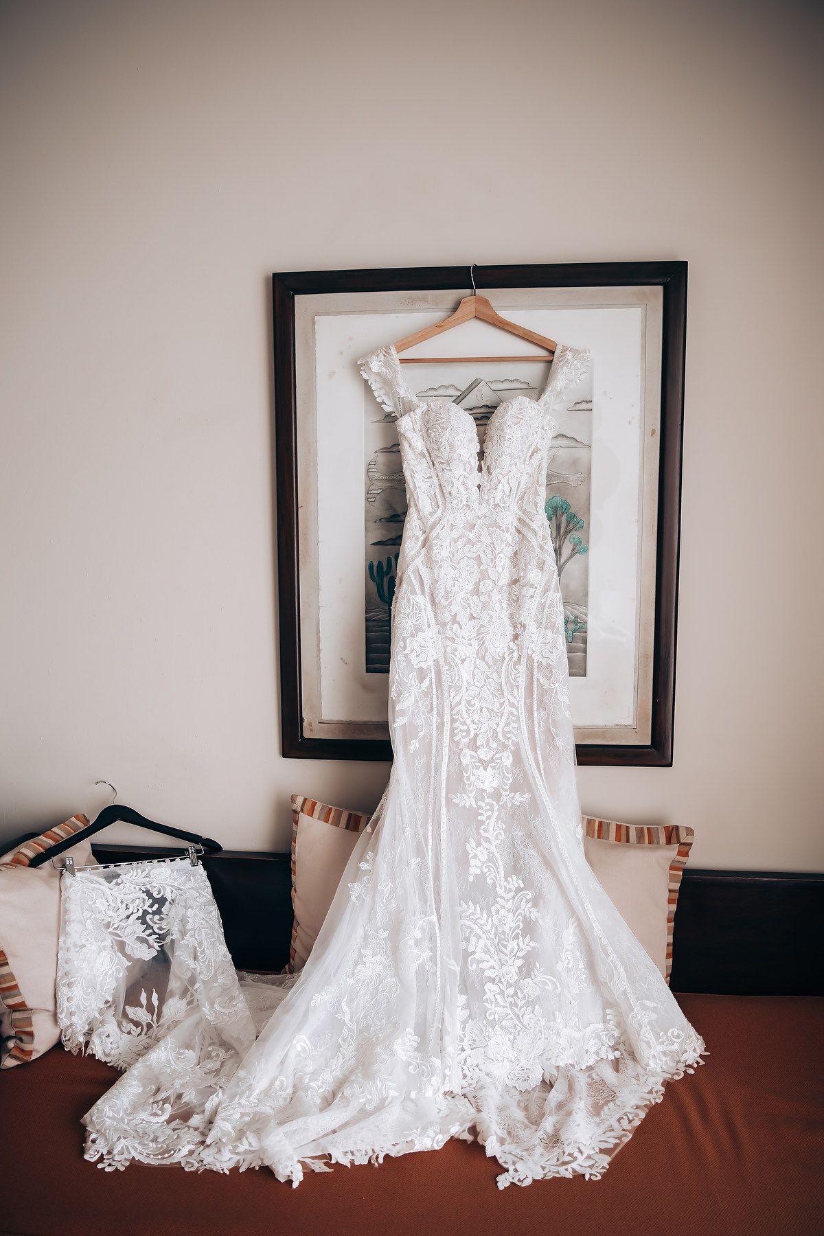 A beautiful white lace wedding dress with intricate details hangs elegantly against a neutral wall.