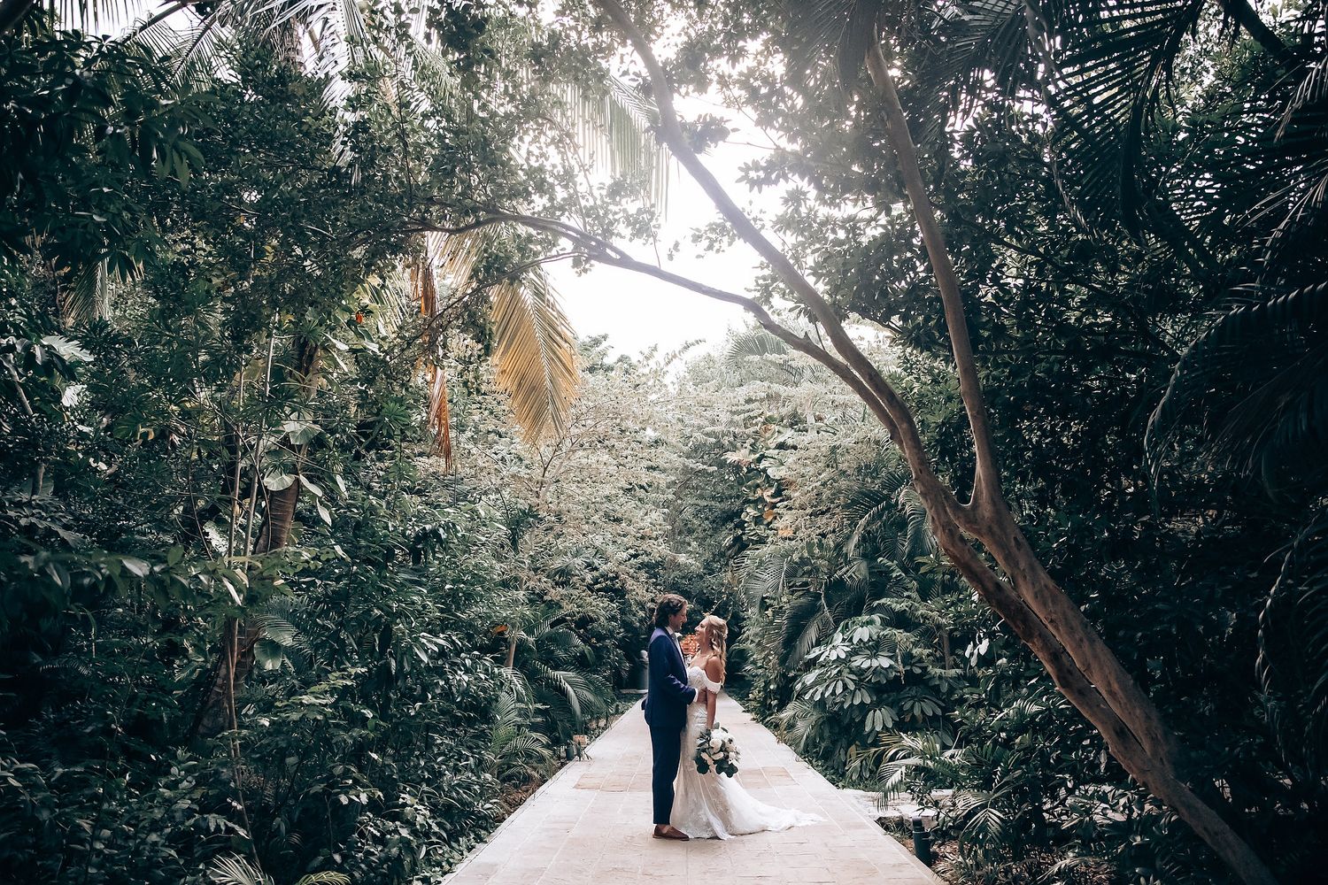 Wedding couple walking through a tree-lined path with natural light filtering through.