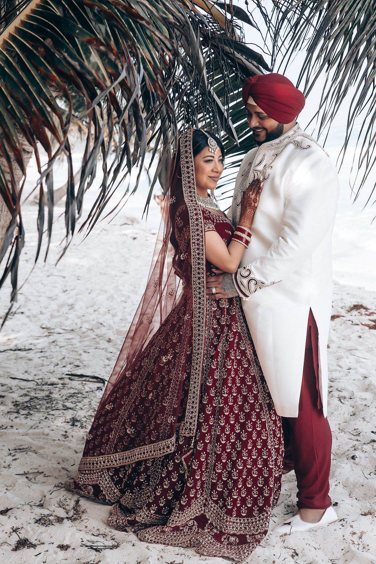 Couple in traditional Indian wedding attire dancing on beach under palm trees.