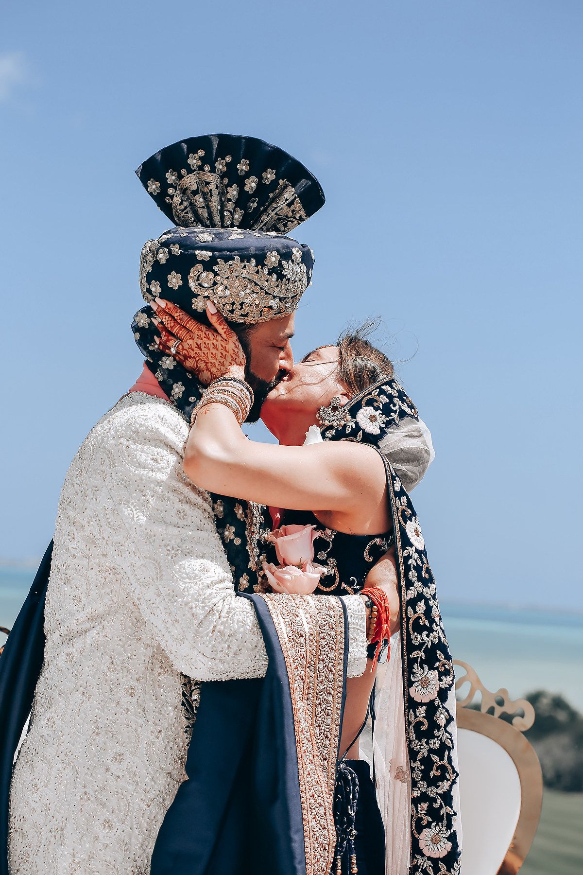 A tender embrace between a couple in traditional Indian wedding attire against a blue sky.