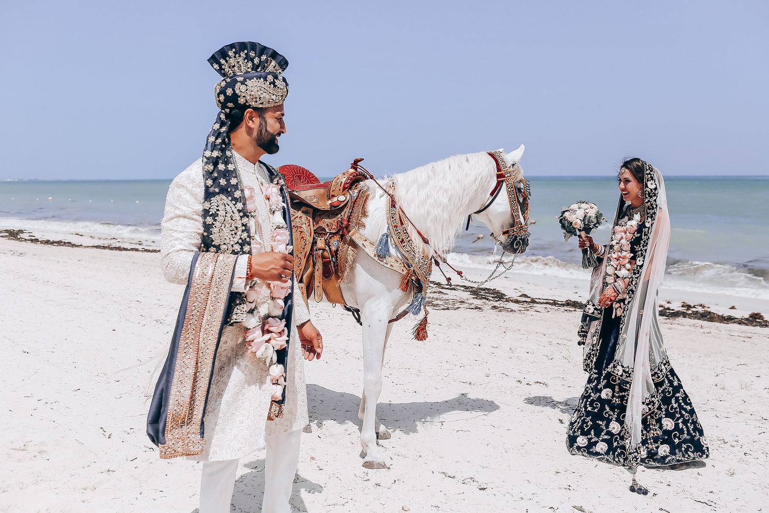 Decorated white horses stand along the shoreline for a beach wedding.