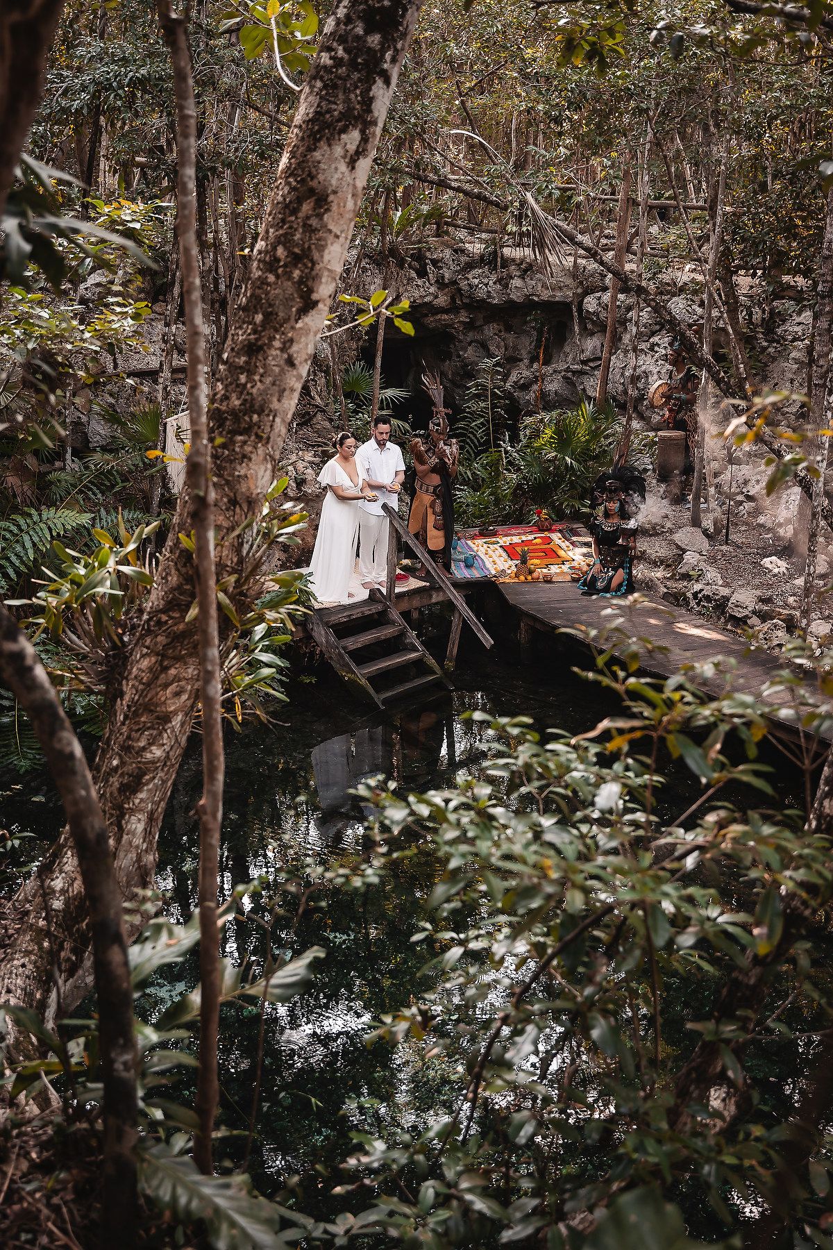A couple sits on wooden steps surrounded by lush tropical foliage and trees in a serene outdoor setting.