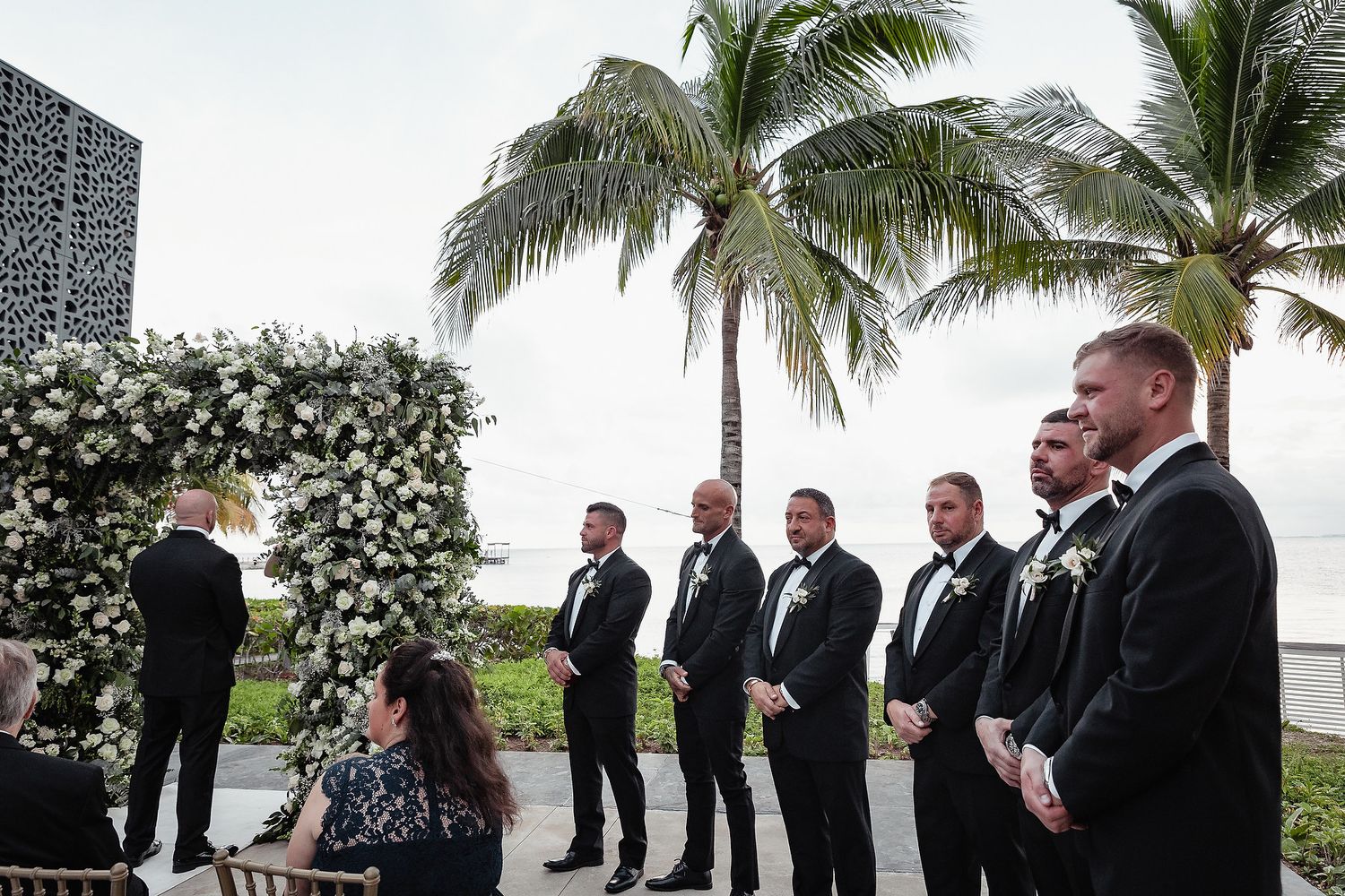 Wedding ceremony on a tropical beach with palm trees and formal wedding party in black suits.