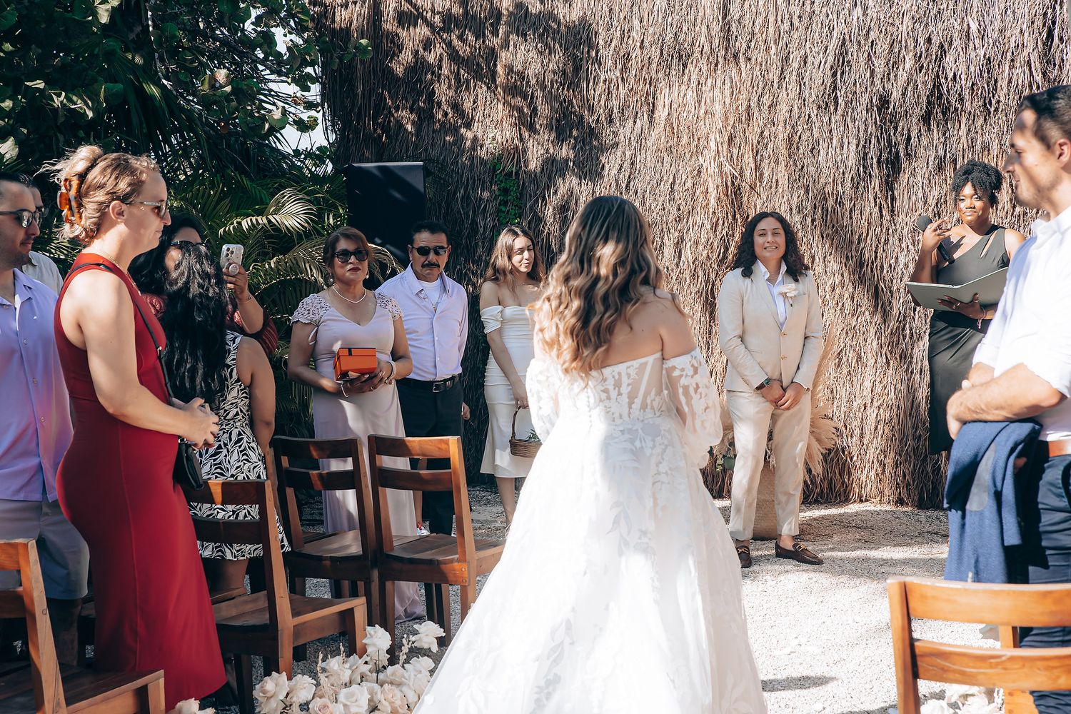 Wedding guests gather outdoors at a tropical venue with rustic wooden chairs and natural decor.