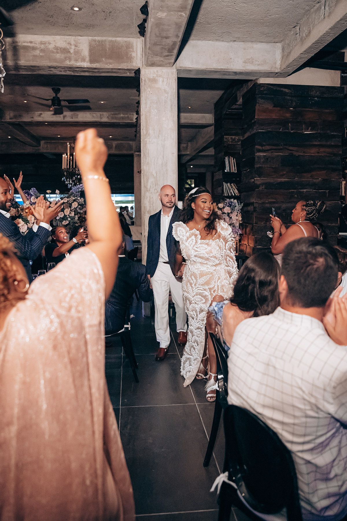 A crowd of wedding guests dancing and celebrating on a dimly lit dance floor at an indoor reception venue.