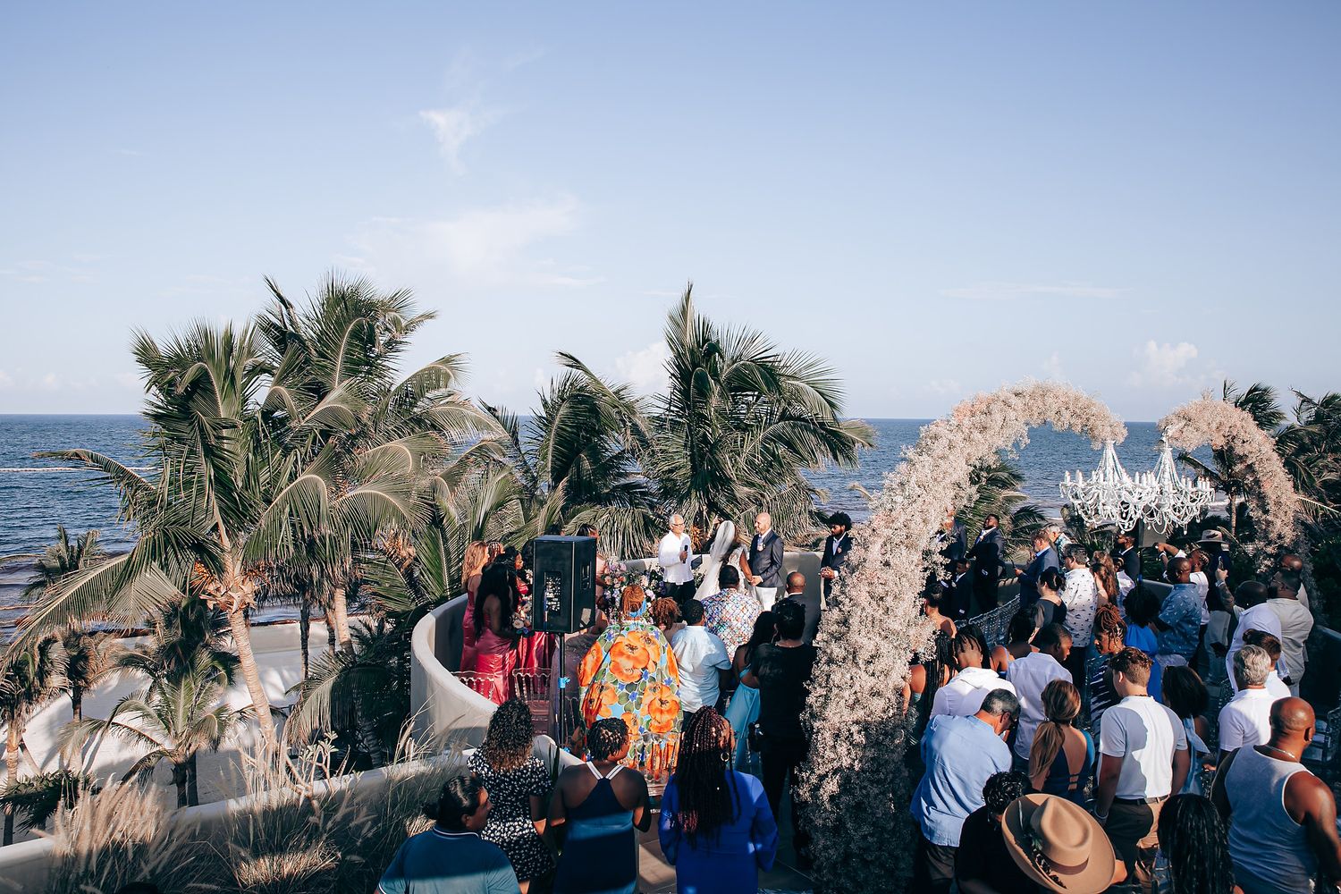 Guests gather at an outdoor beach wedding ceremony under a natural woven arch with palm trees swaying in the breeze.