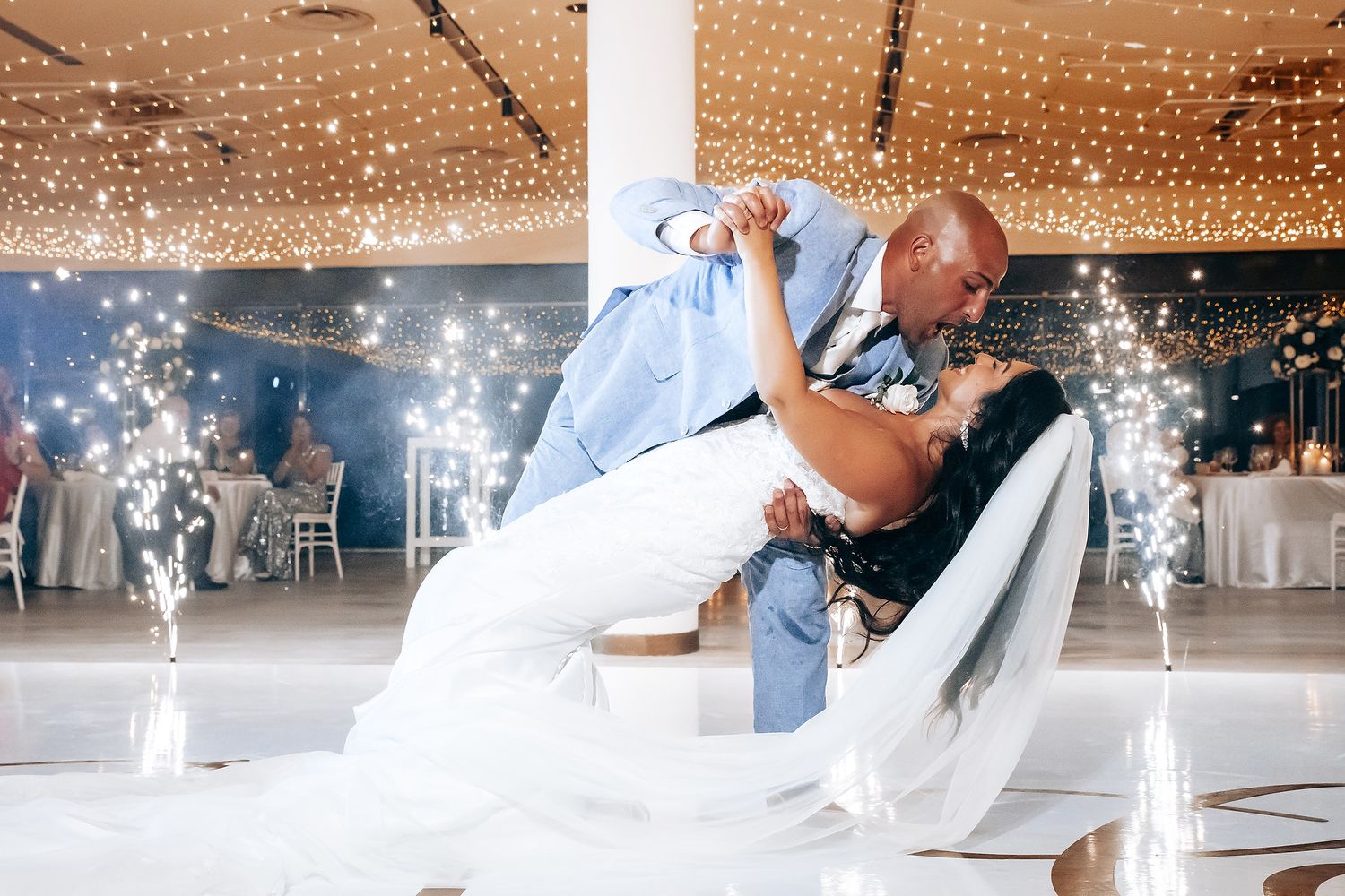 A couple shares a romantic dance on a white dance floor under twinkling string lights at an elegant reception venue.