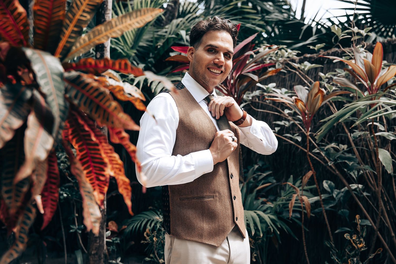 Wedding guests mingling outdoors among tropical plants and palm fronds at garden reception.