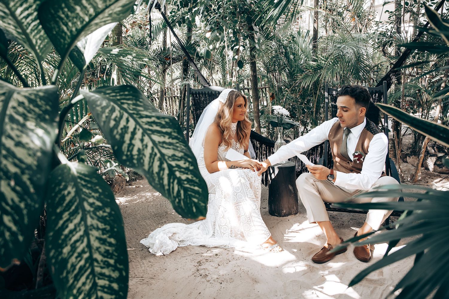 Couple in wedding attire sits surrounded by lush tropical foliage during an intimate outdoor ceremony.