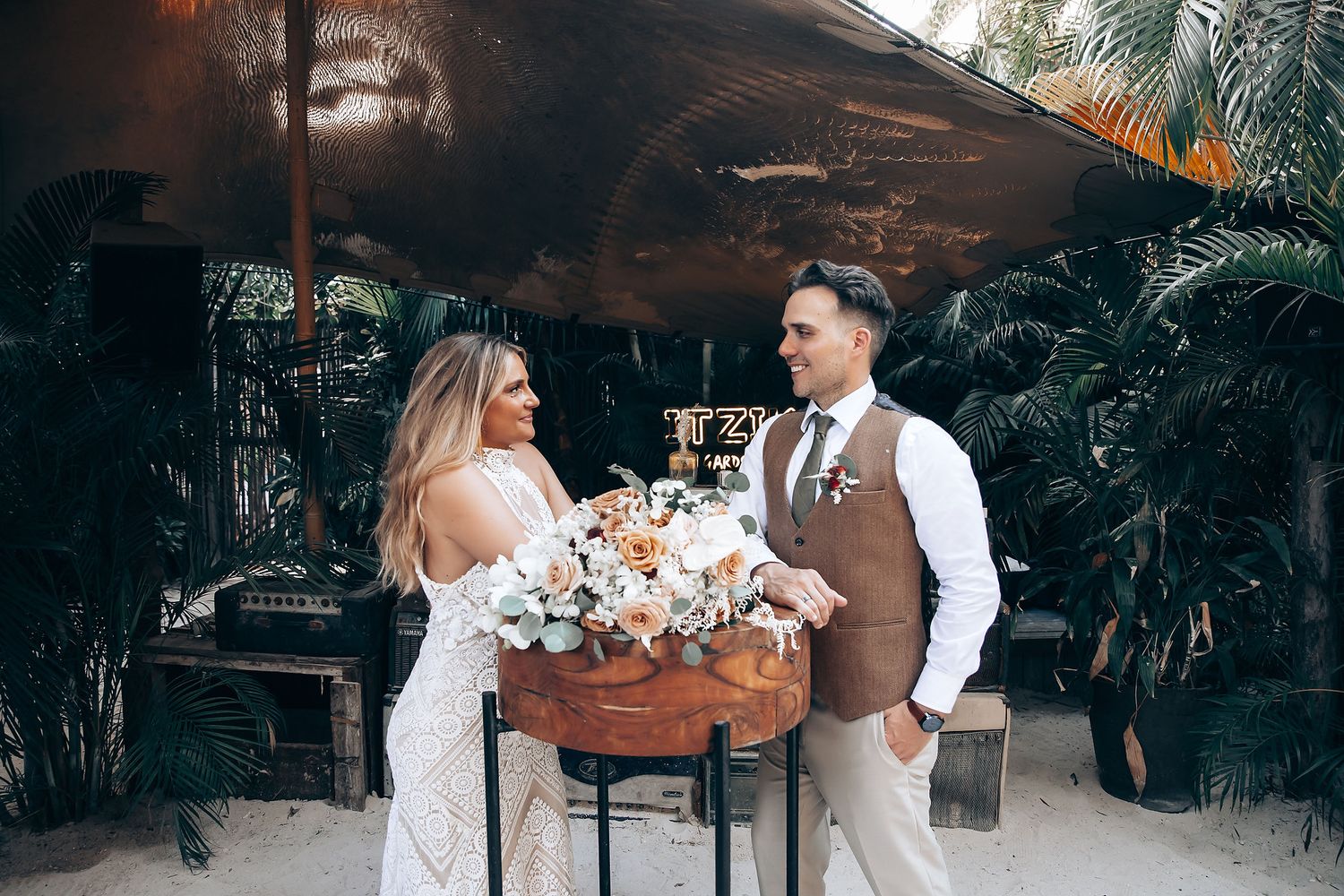 A romantic tropical wedding scene with a couple standing beside a floral arrangement in a lush garden setting.