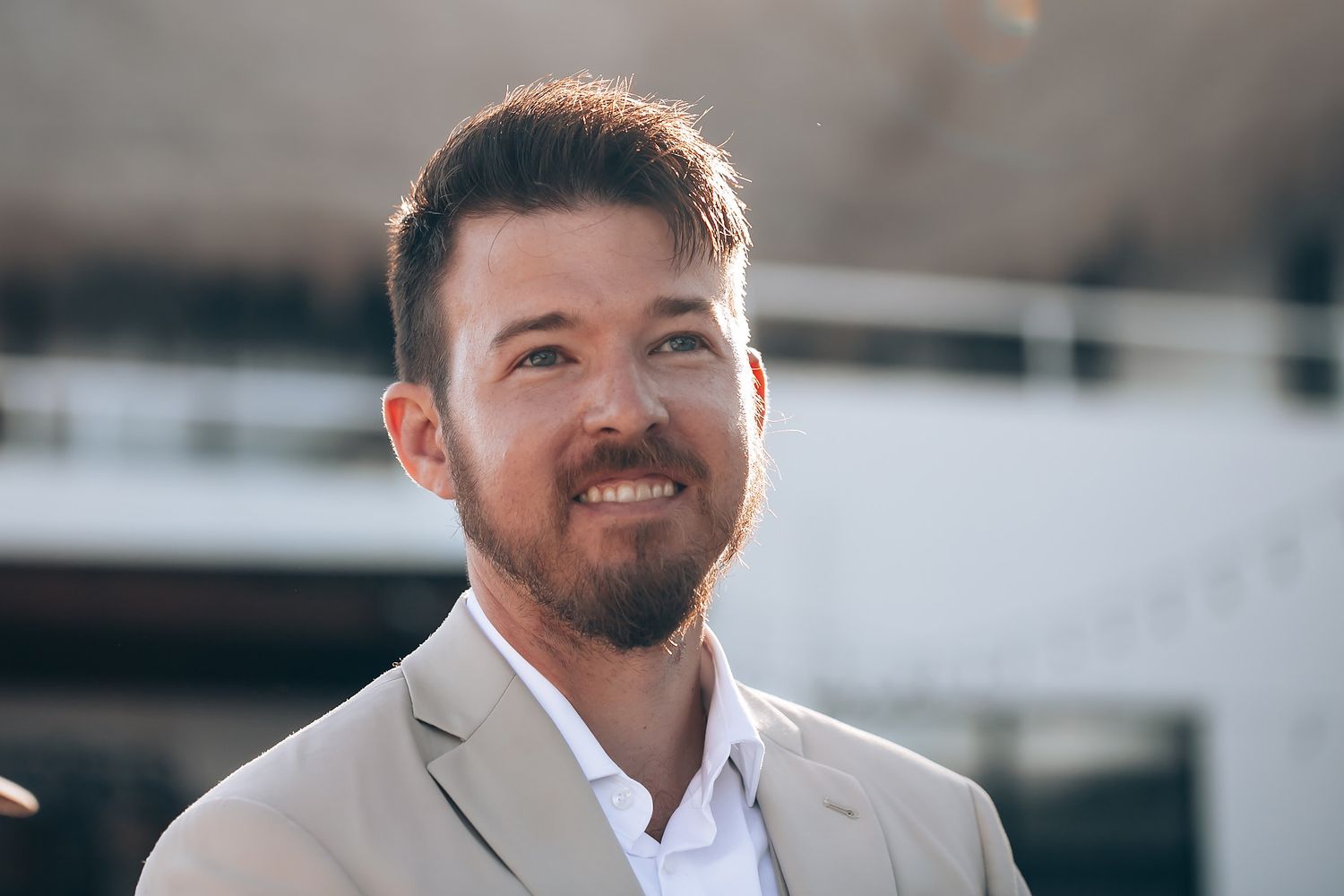 Groom in light colored suit smiles warmly in outdoor wedding venue setting.