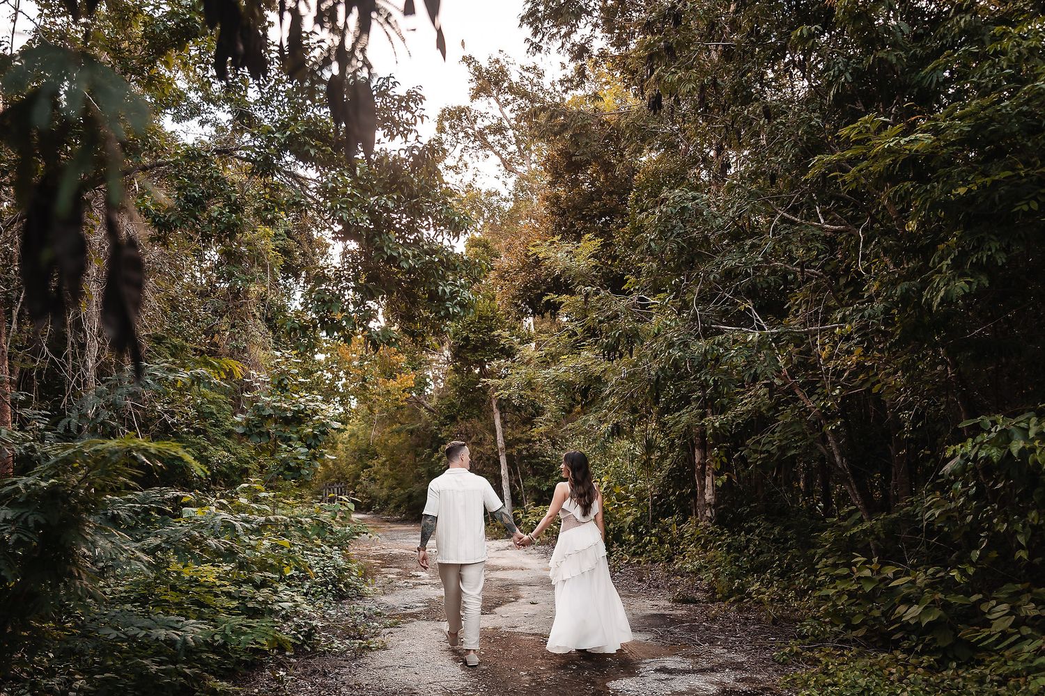 Two people in elegant white outfits walk along a scenic forest trail during sunset.