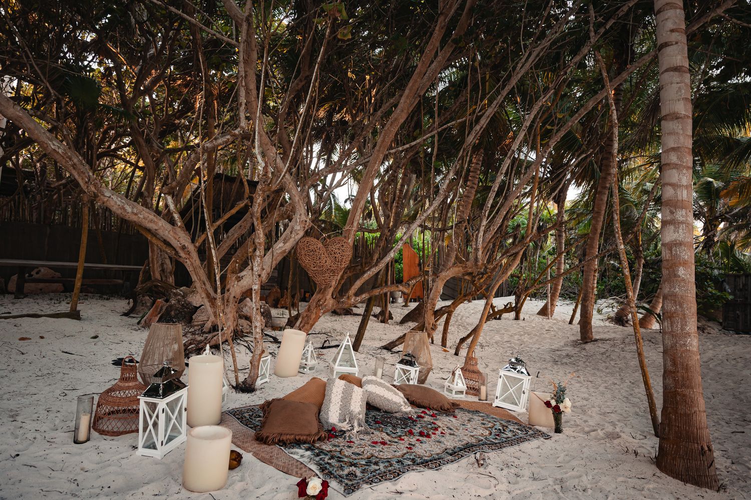 Cozy beach lounge area with lanterns and rugs nestled between tropical trees.
