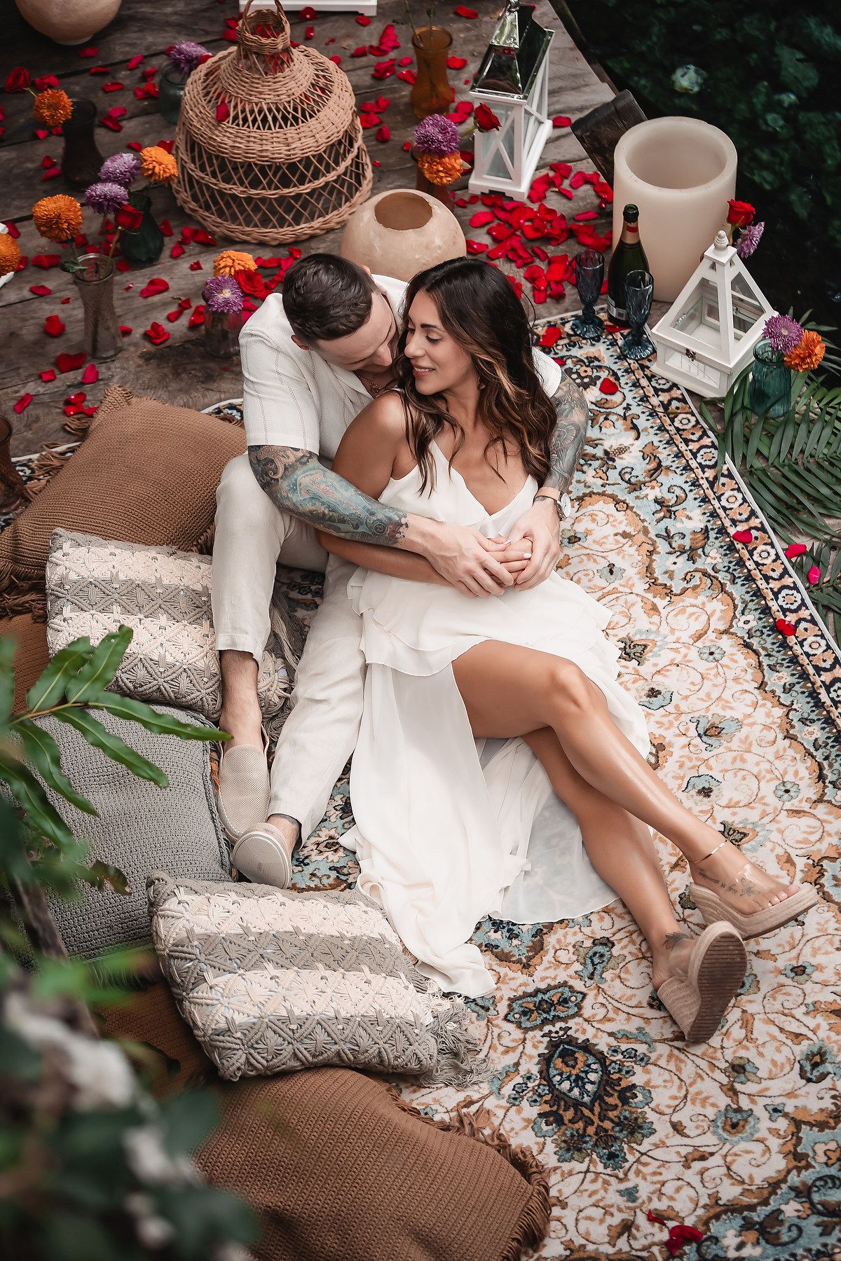 Romantic outdoor setup with white lanterns, rose petals, and bohemian decor on patterned rug.