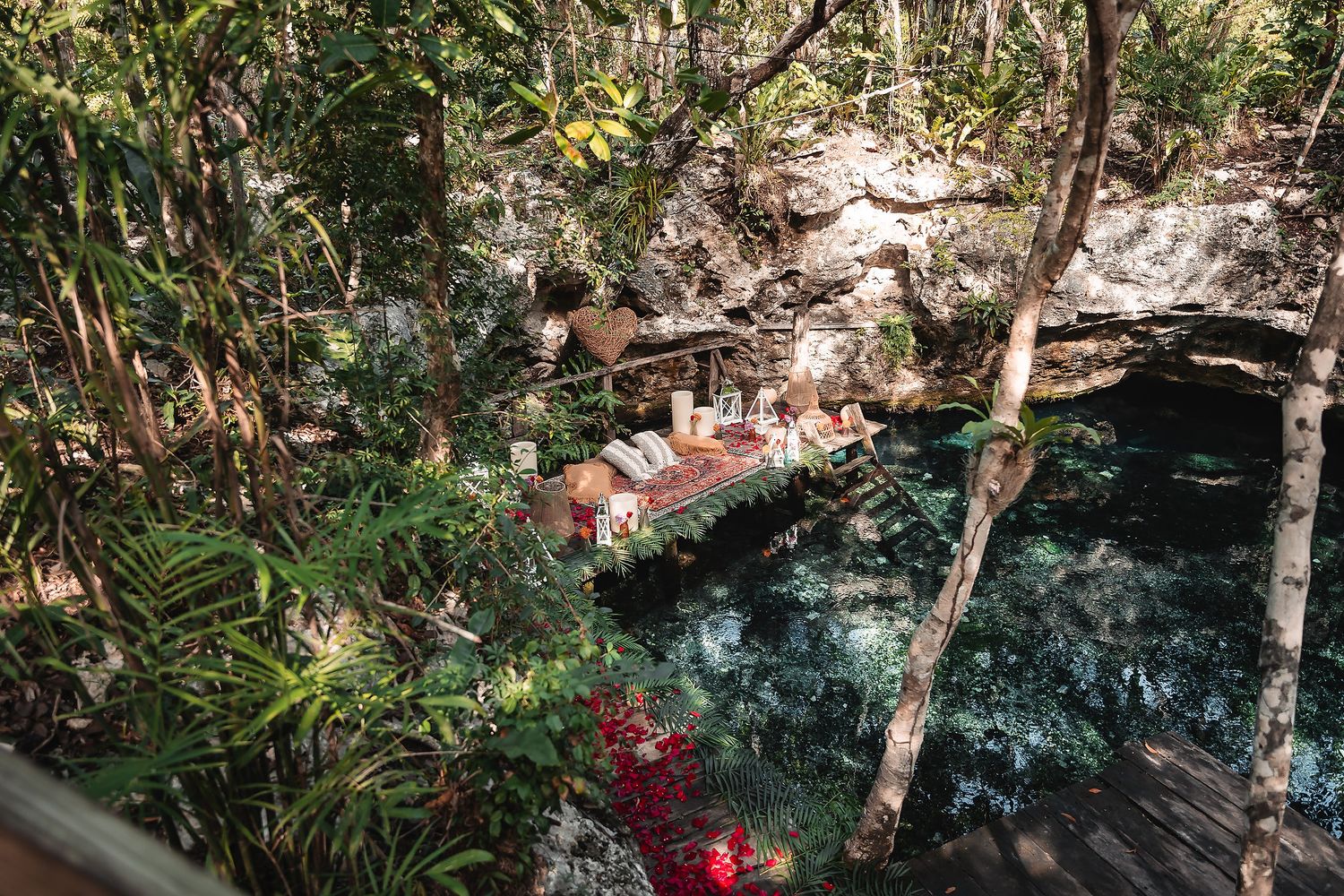 Romantic picnic setup with red rose petals along the edge of a crystal clear cenote.