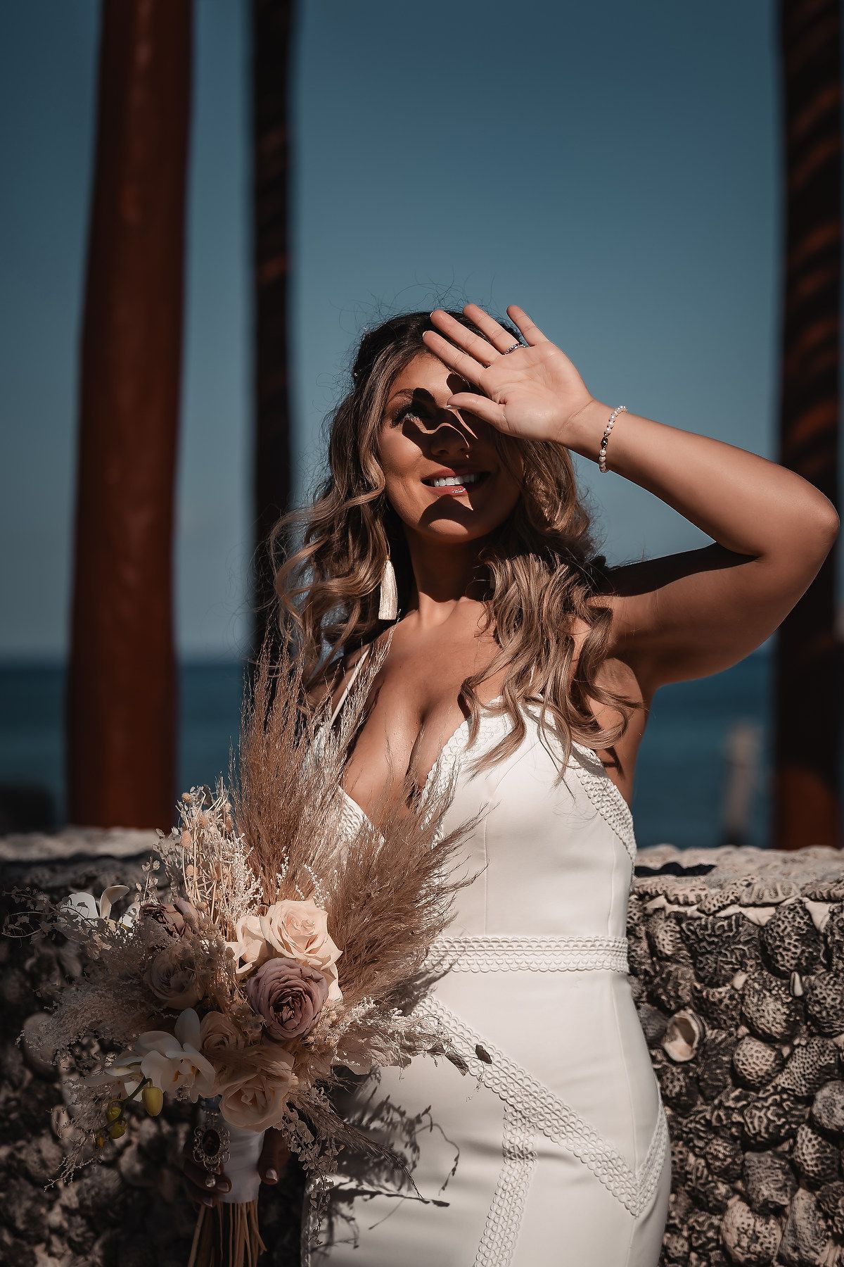 A bride in a white dress poses dramatically against a stone wall with dried pampas grass decorations.
