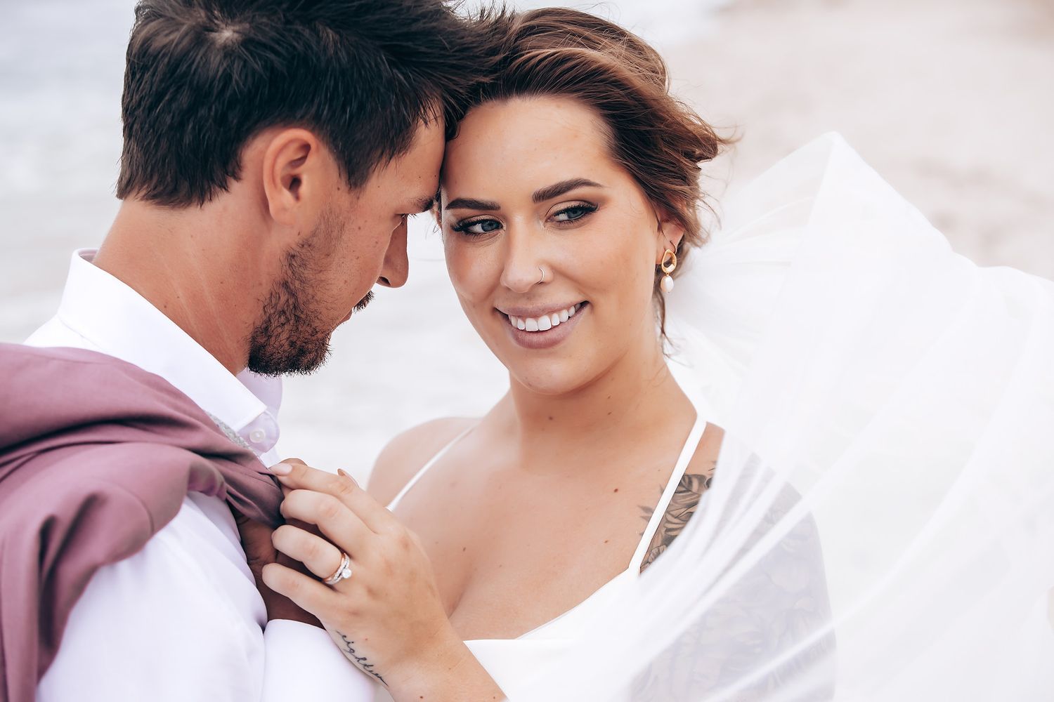A romantic beach wedding photoshoot with flowing veil and soft intimate poses against an overcast sky.