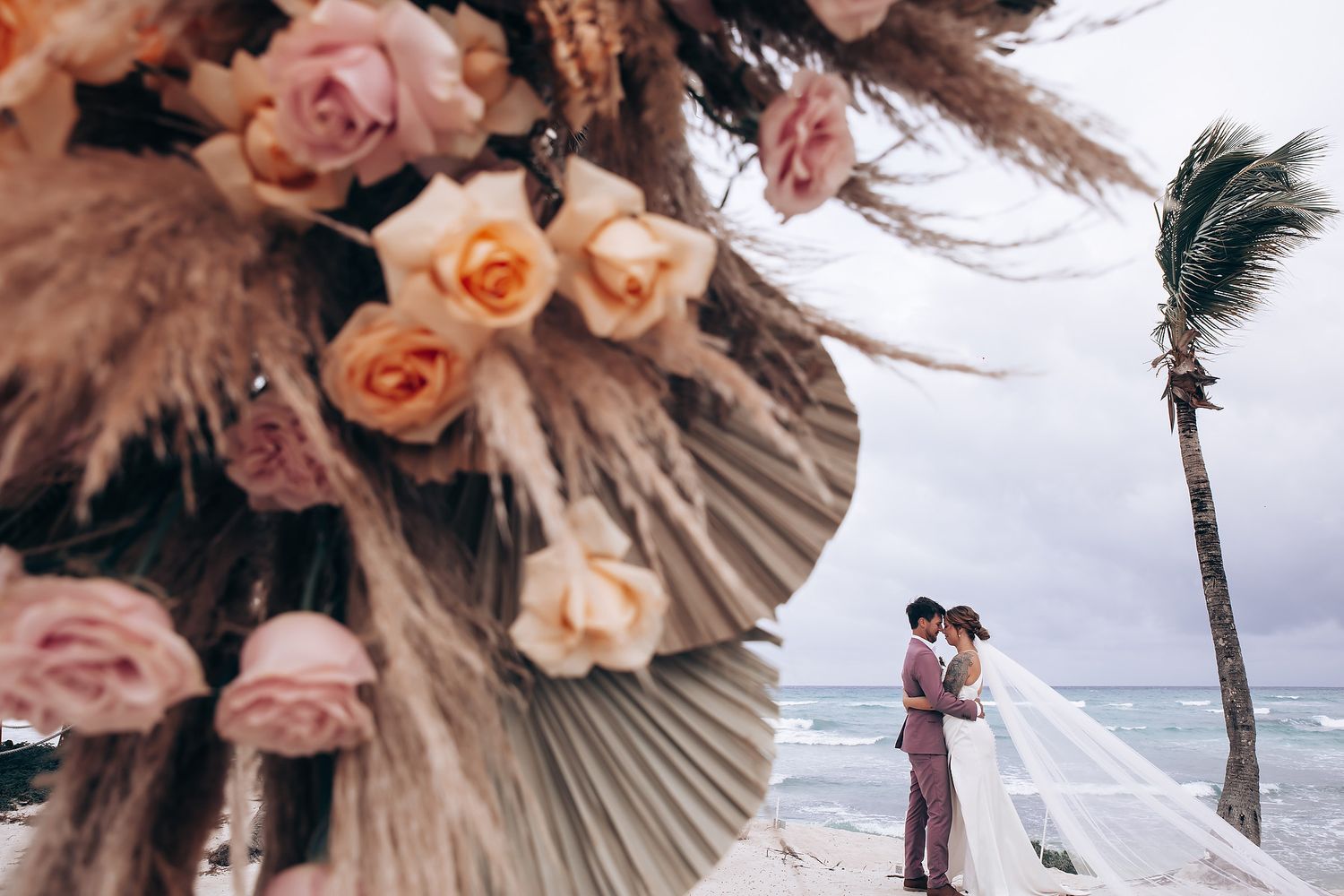 Elegant peach and blush floral arrangements with palm fronds decorate a beachside wedding setting.