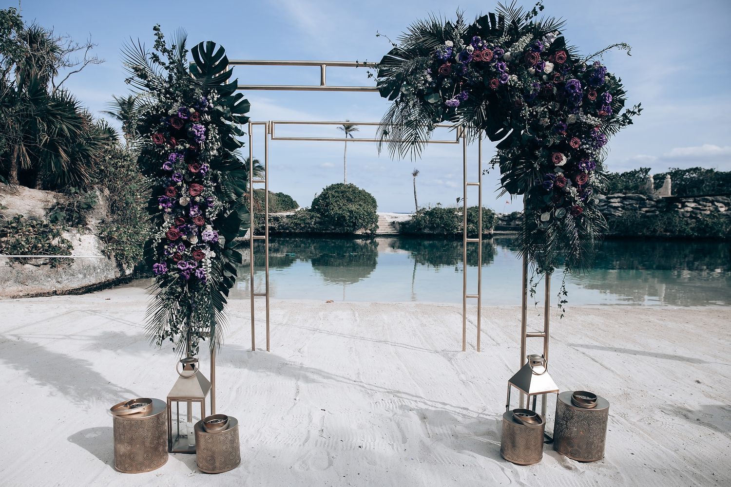 Floral wedding arch setup with lanterns on a tropical beach during sunset.